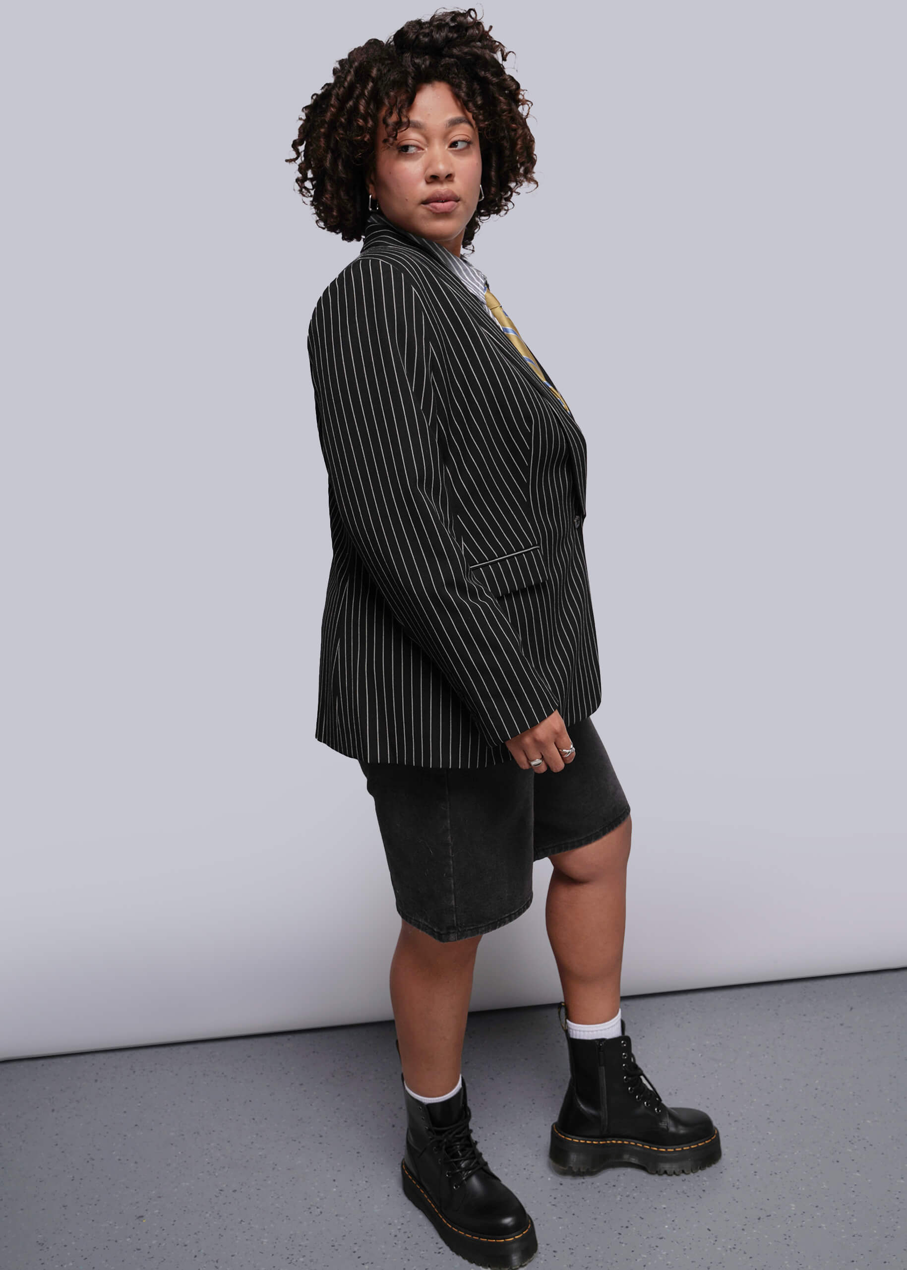 1X=Person wearing a black pinstripe blazer, black shorts, and black boots against a white background