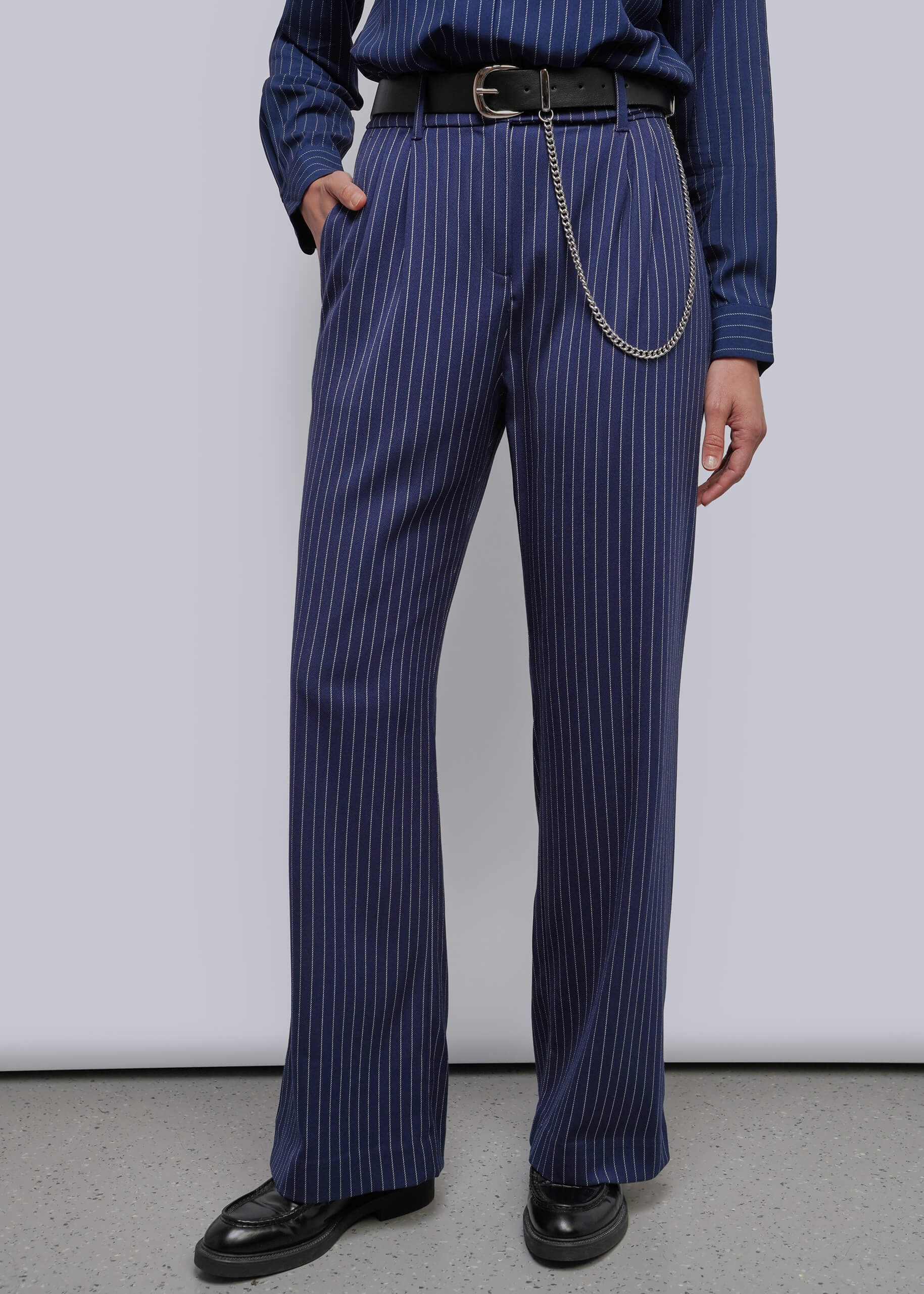 S=A person wearing navy blue pinstriped trousers with a black belt and silver chain detail, paired with a matching navy shirt and black shoes, photographed against a plain light gray studio background.