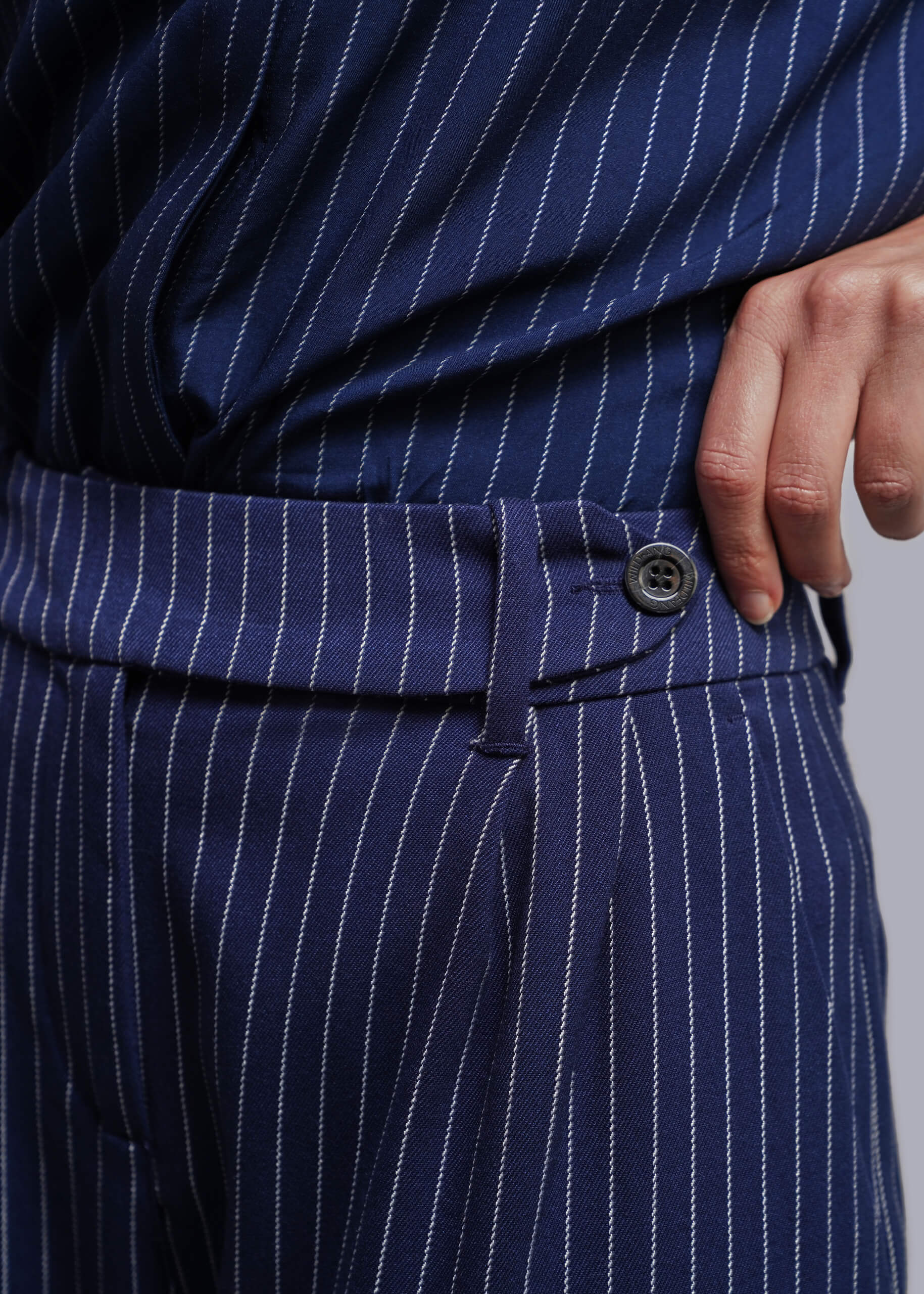Close-up of navy blue pinstriped trousers with a matching top, showing the waistband, button, and a hand resting near the pocket.
