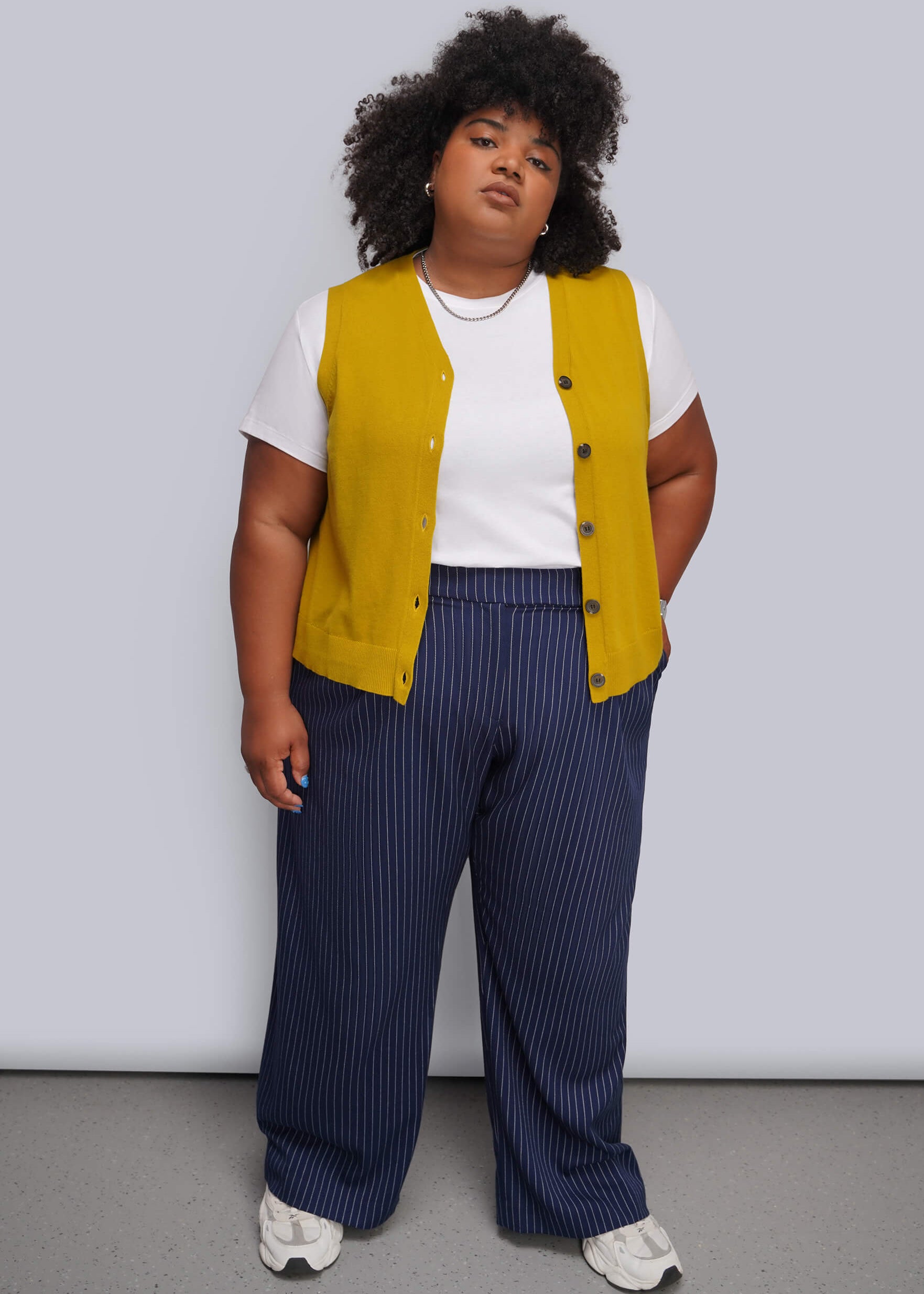 1X=A plus-size model stands against a light gray studio background wearing a mustard yellow sleeveless vest over a white t-shirt, paired with navy blue pinstripe trousers and white sneakers.