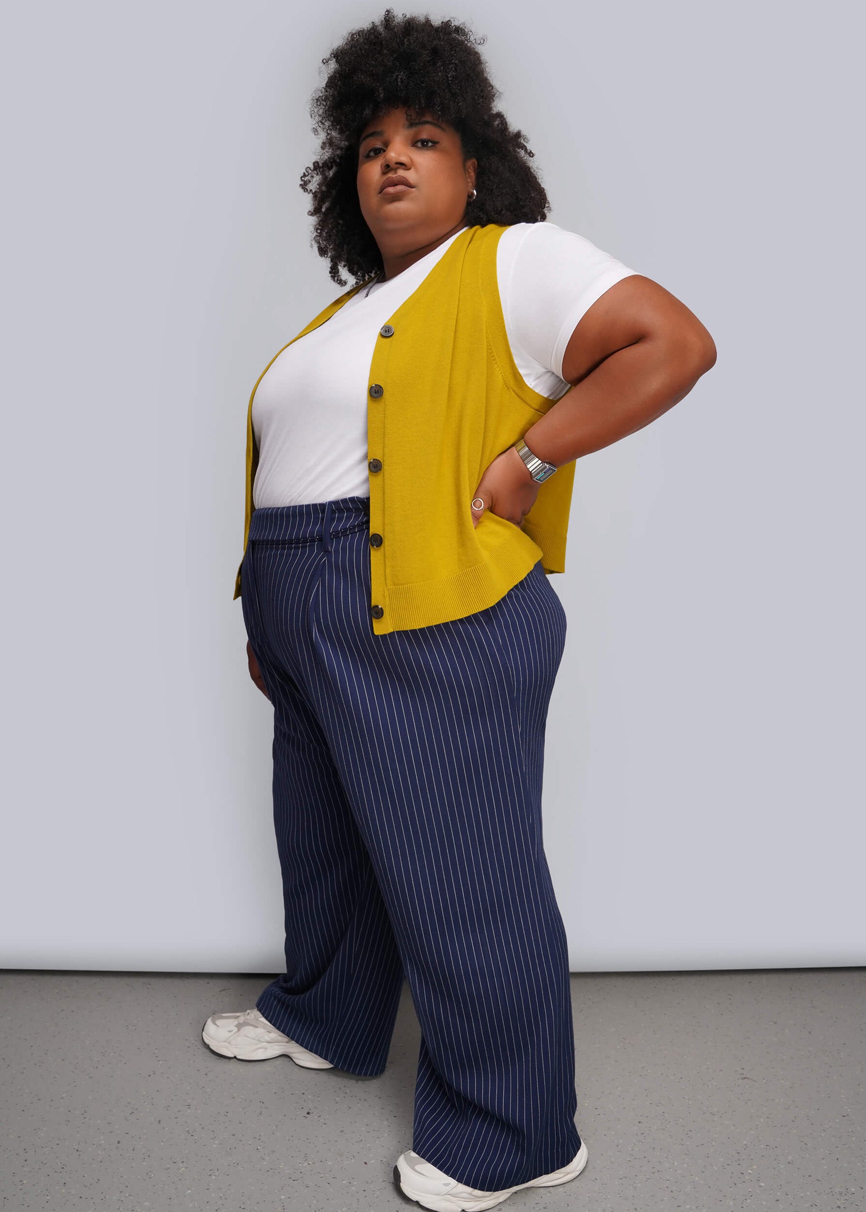 1X=A plus-size model stands in a studio wearing a white t-shirt, a mustard yellow button-front vest, navy blue pinstriped wide-leg trousers, and white sneakers against a light gray background.