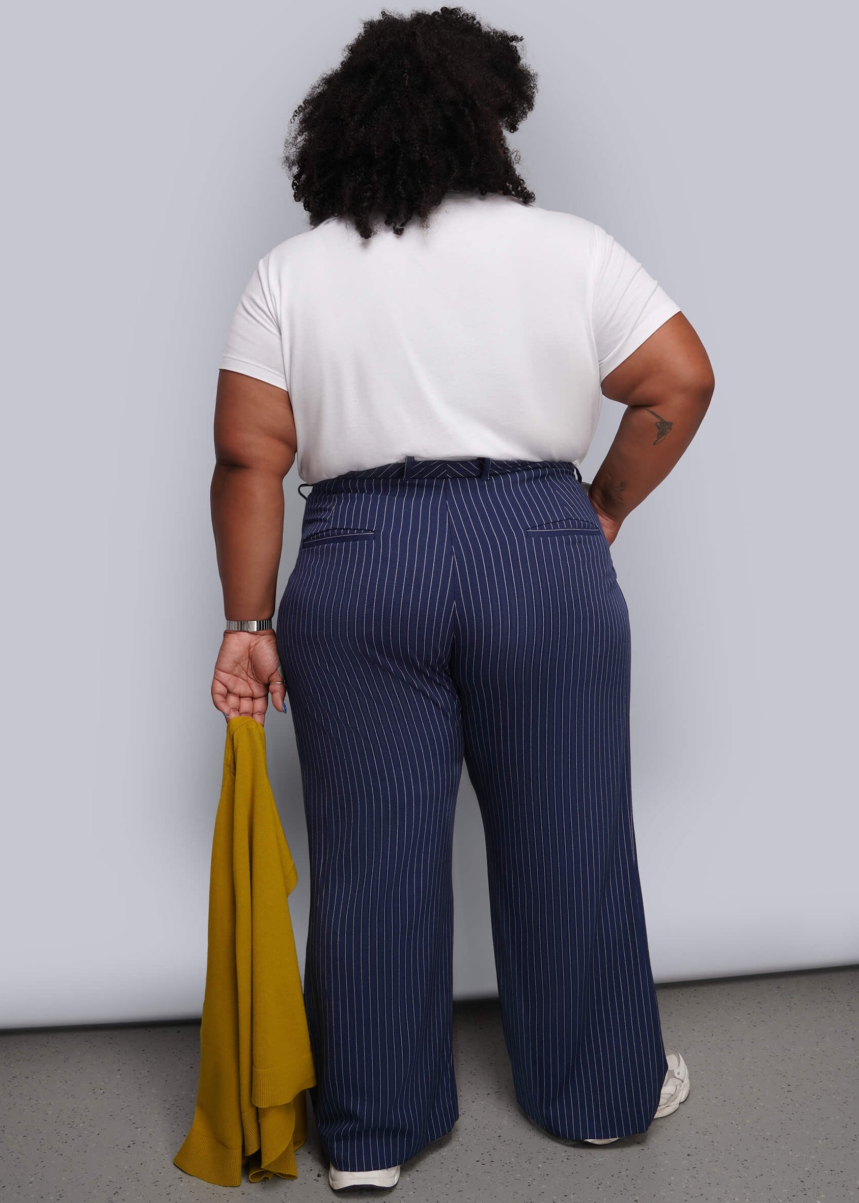 1X=A person shown from the back wearing navy blue pinstripe wide-leg trousers, a white t-shirt, and white sneakers, holding a mustard yellow garment against a plain light gray wall.