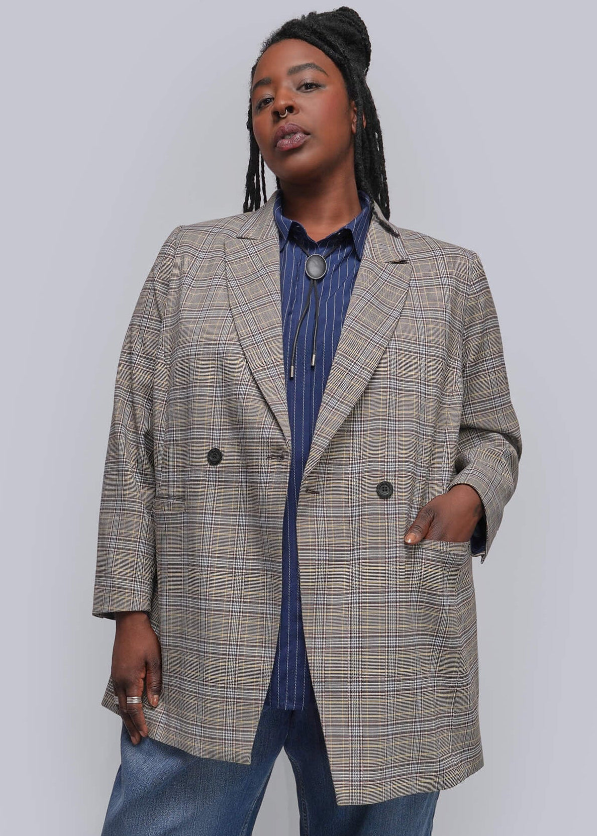 1X=A model wearing an oversized beige and brown plaid blazer over a blue striped button-down shirt, paired with cuffed blue jeans and black pointed-toe ankle boots, standing against a plain light gray studio background.