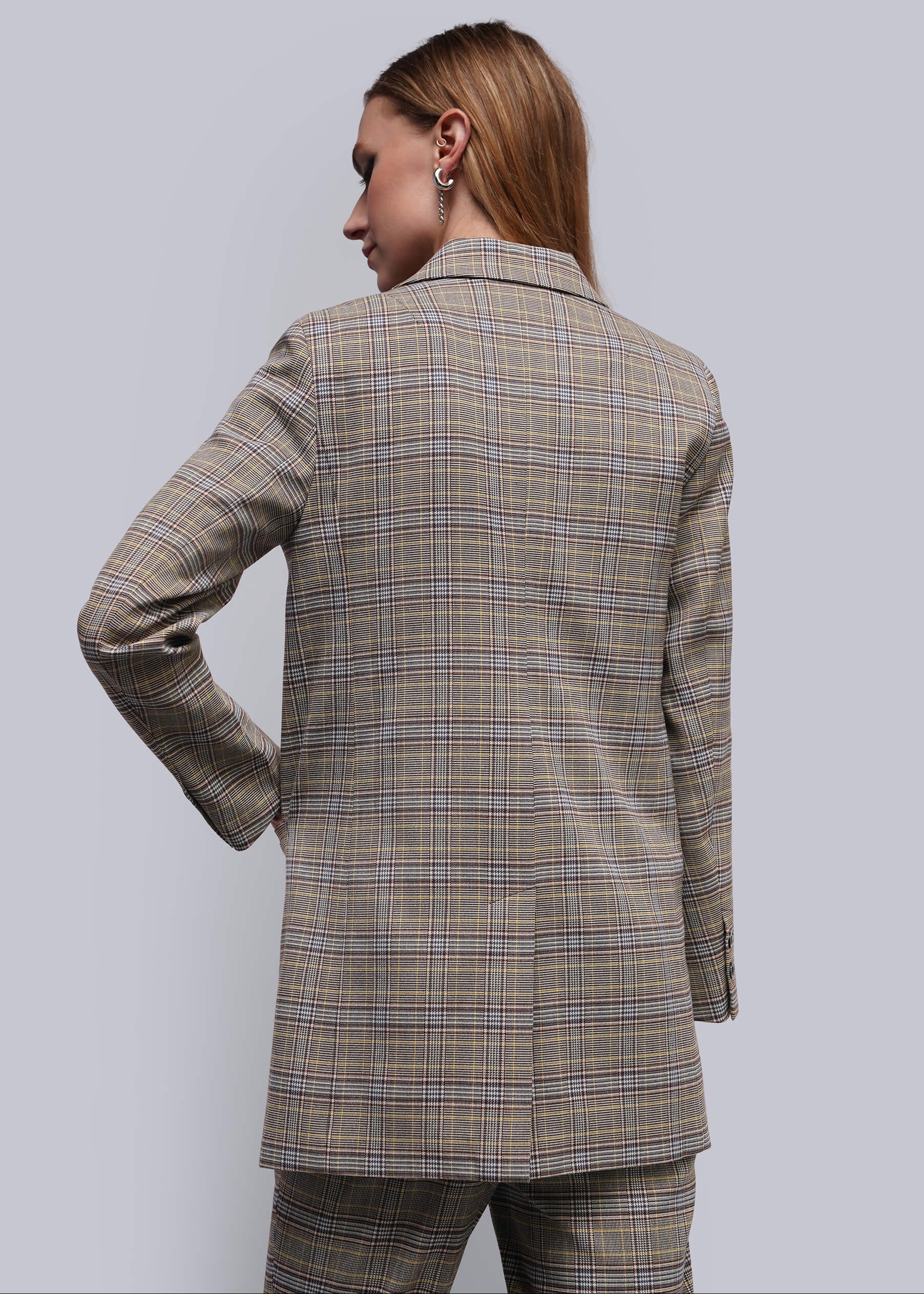 S=A woman shown from the back wearing a beige plaid blazer and matching trousers against a solid gray studio background.