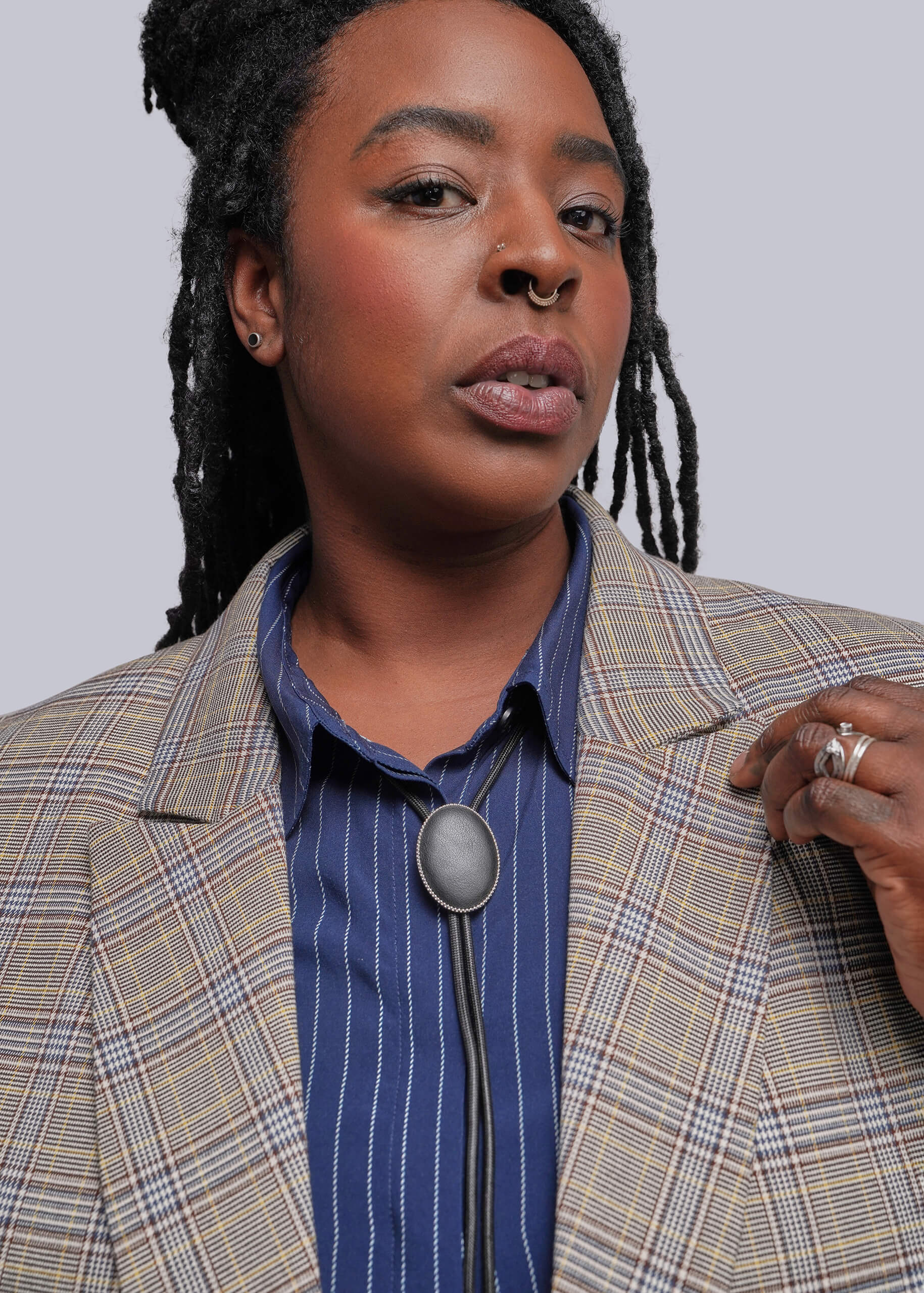 1X=A close-up portrait of a person wearing a beige plaid blazer over a blue striped button-down shirt with a large oval bolo tie pendant.