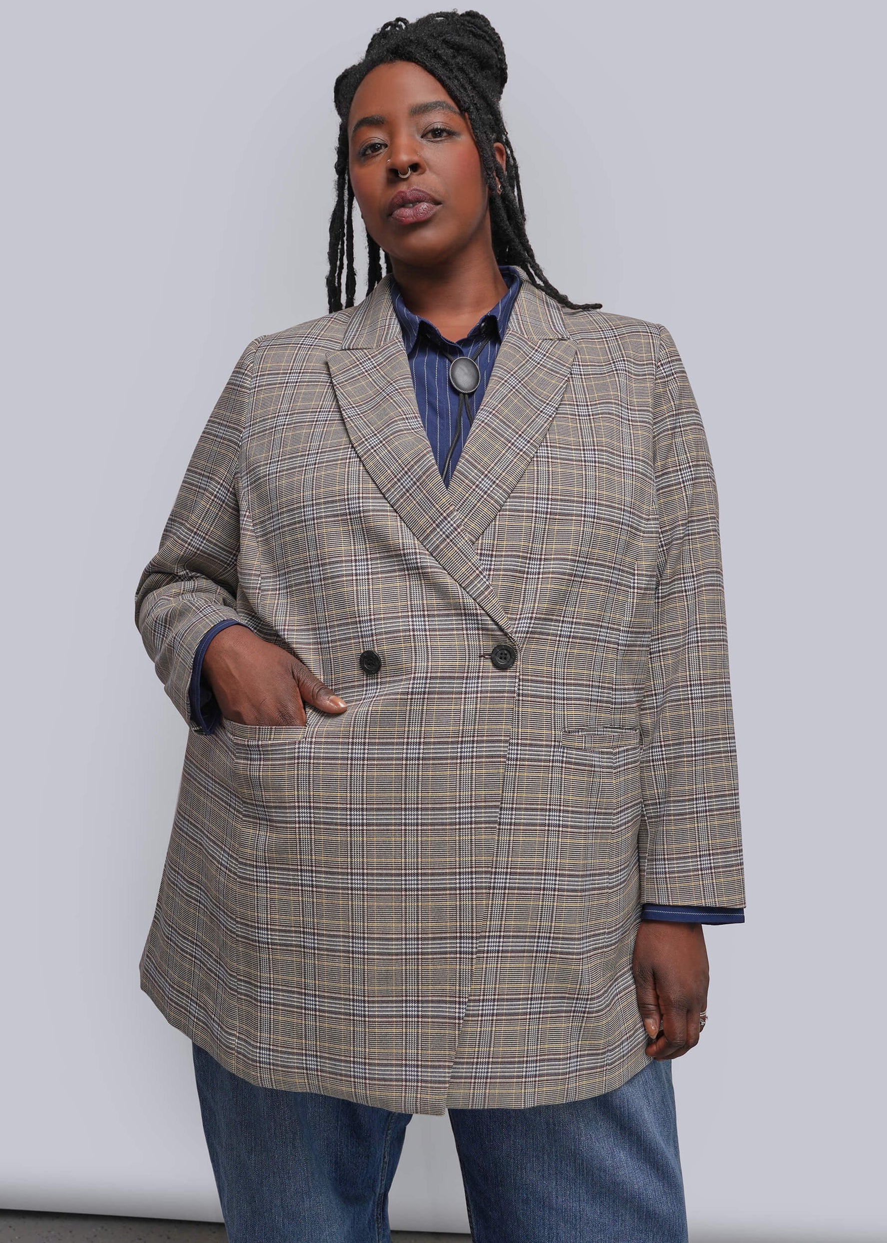 1X=A model wearing a beige and brown plaid double-breasted blazer over a blue collared shirt and blue jeans, standing against a light gray studio background.