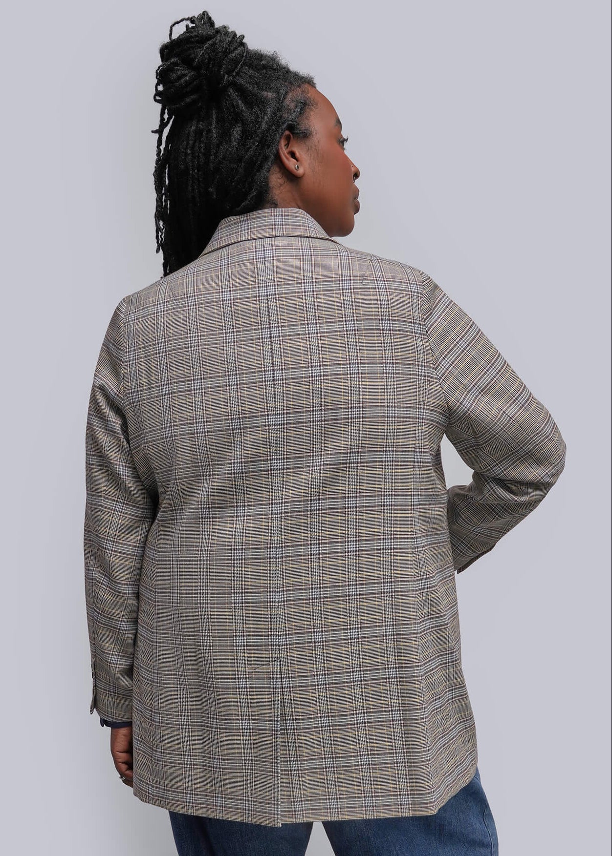 1X=A woman shown from the back wearing a beige and brown plaid blazer, blue jeans, and black pointed-toe heels against a plain light gray studio background.