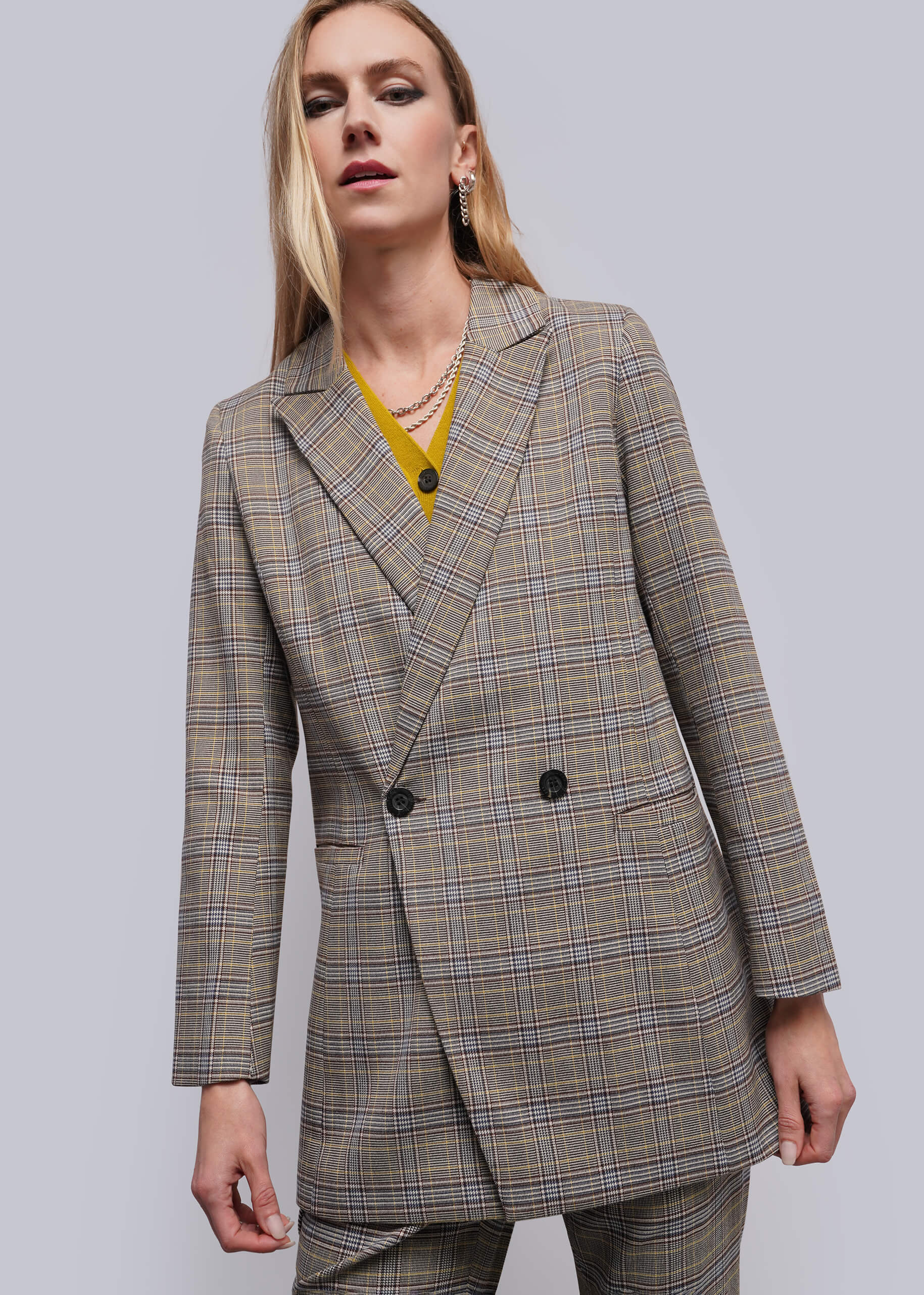 S=A woman wearing a double-breasted plaid blazer in beige, brown, and gray tones, styled with a yellow top and layered necklaces, against a solid gray studio background.