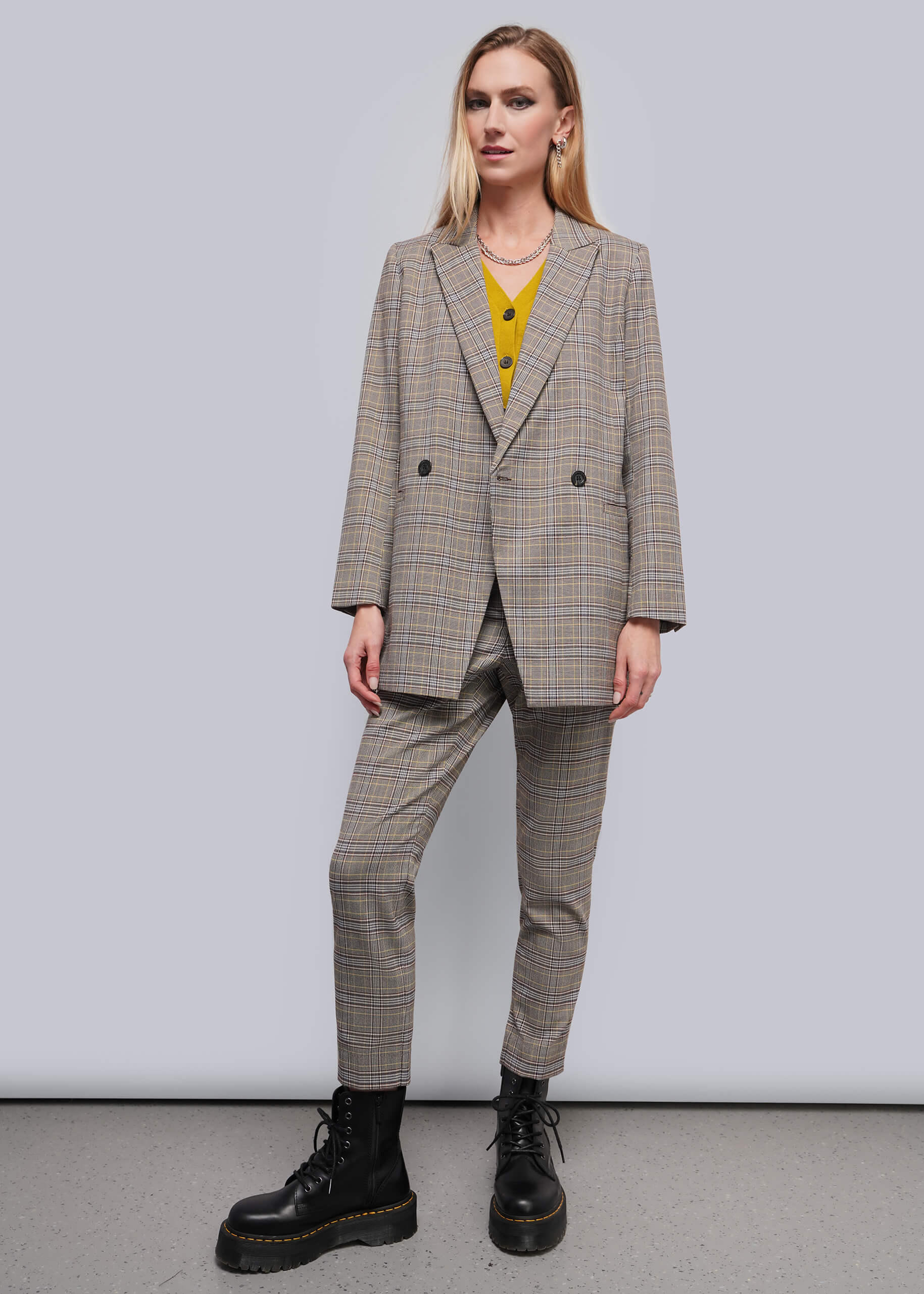 S=A woman modeling a beige and brown plaid double-breasted blazer with matching trousers, worn over a yellow top and paired with black combat boots, against a plain gray studio background.