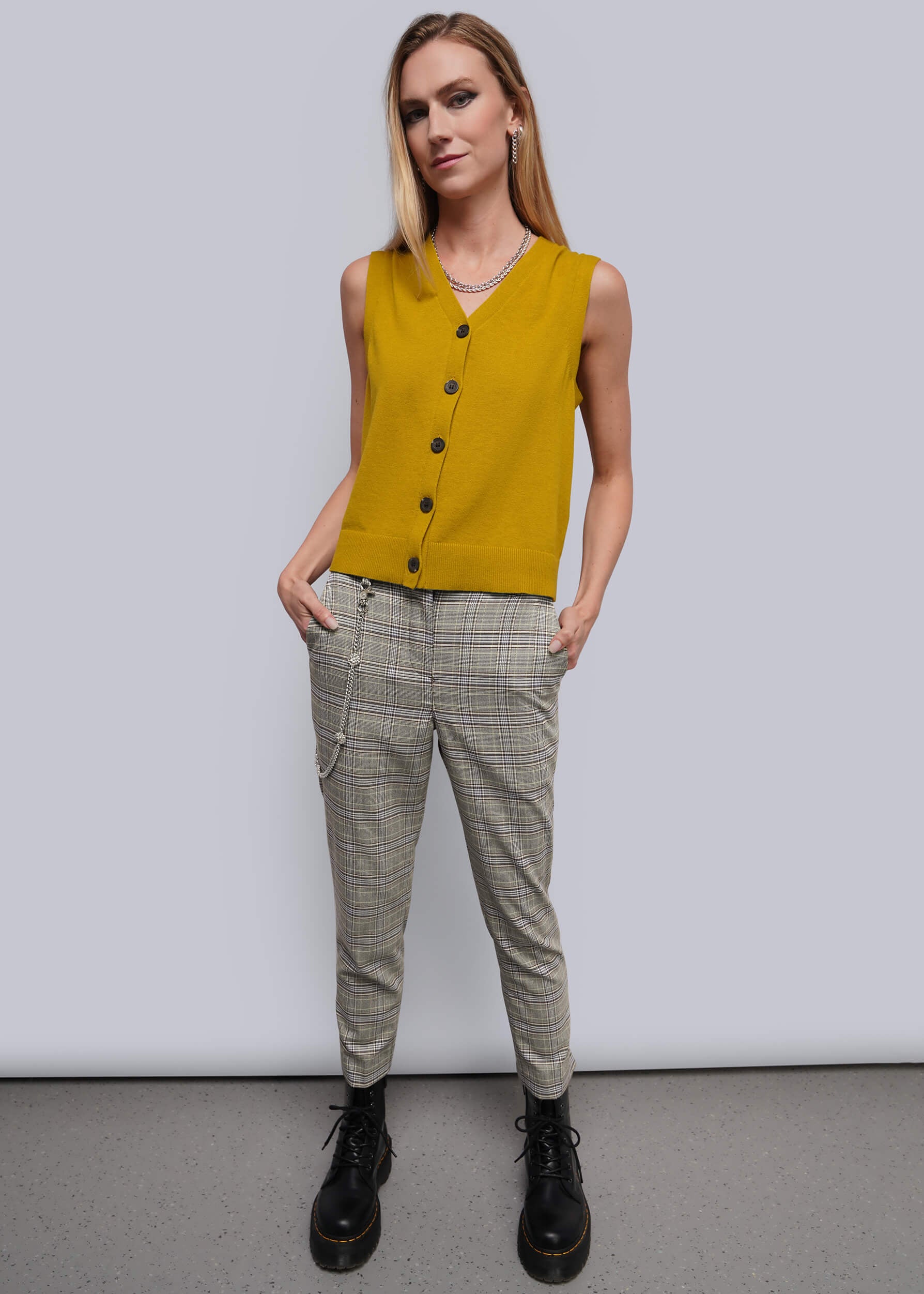 A woman modeling a sleeveless mustard yellow knit vest with black buttons, paired with beige plaid trousers and black boots, standing against a light gray studio background.