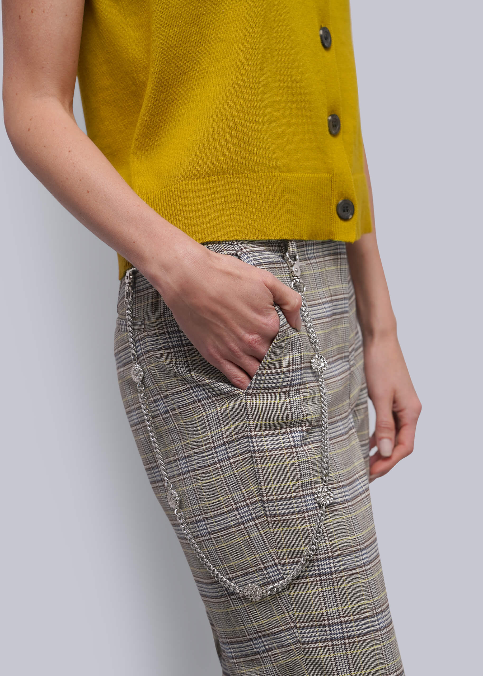 A close-up of a person wearing a mustard yellow sleeveless cardigan and beige plaid pants with silver chain details, against a light gray background.