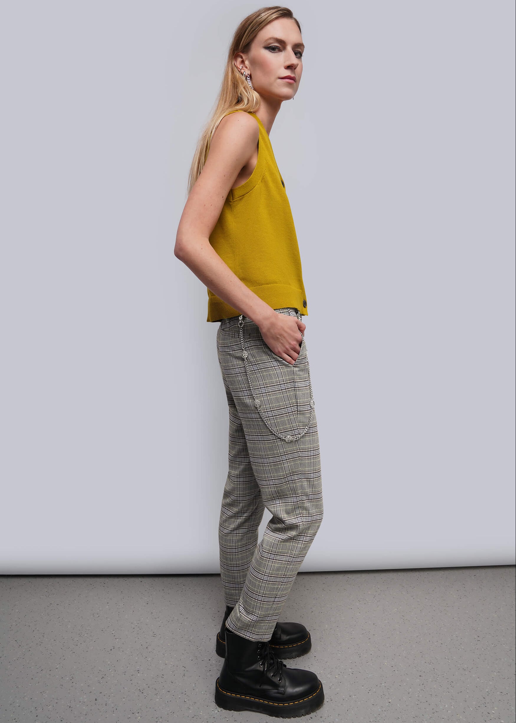 S=A woman modeling a sleeveless mustard yellow top and gray plaid trousers with black boots, standing against a light gray studio background.