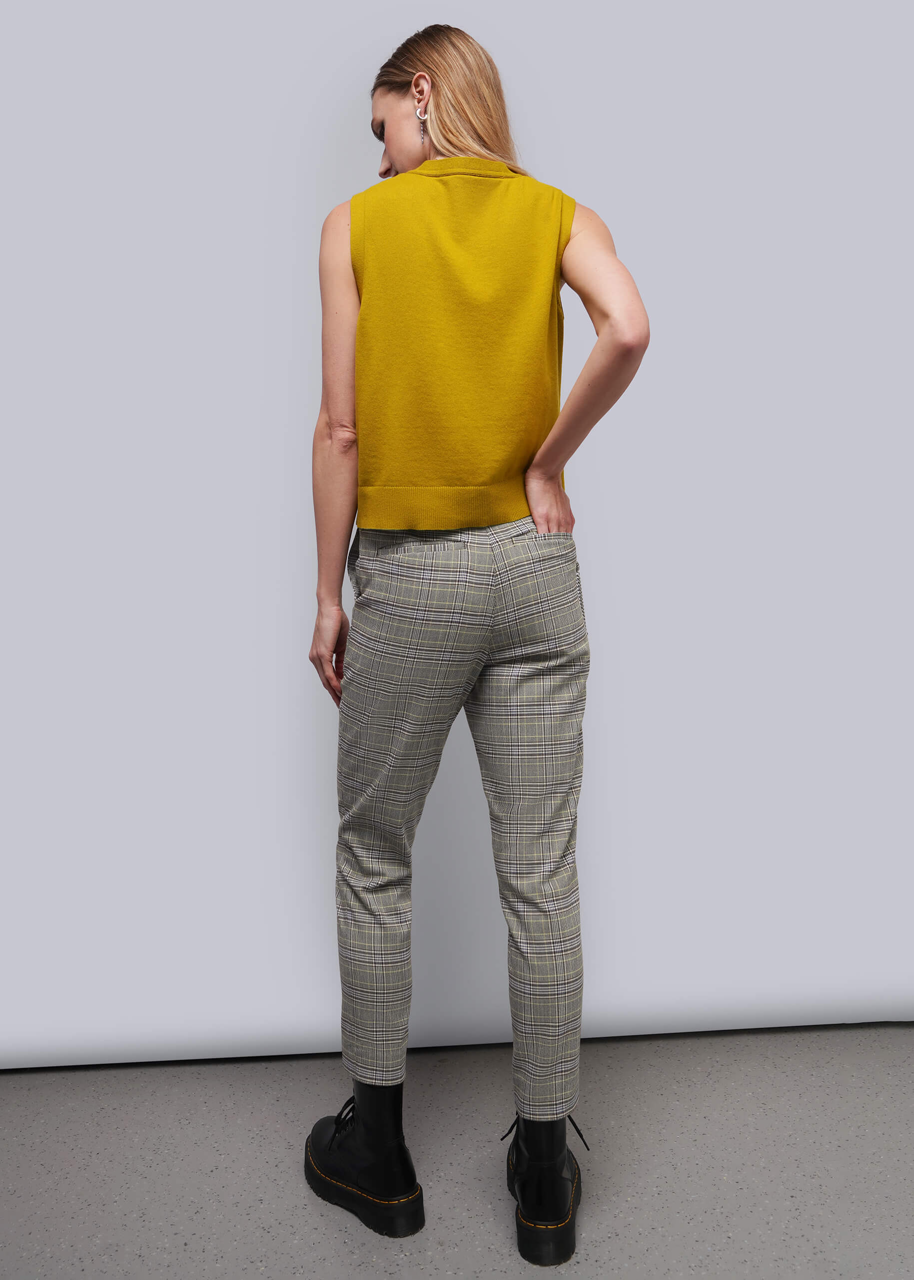 S=A woman shown from behind wearing a sleeveless mustard yellow knit top, gray and beige plaid trousers, and black boots against a light gray studio background.