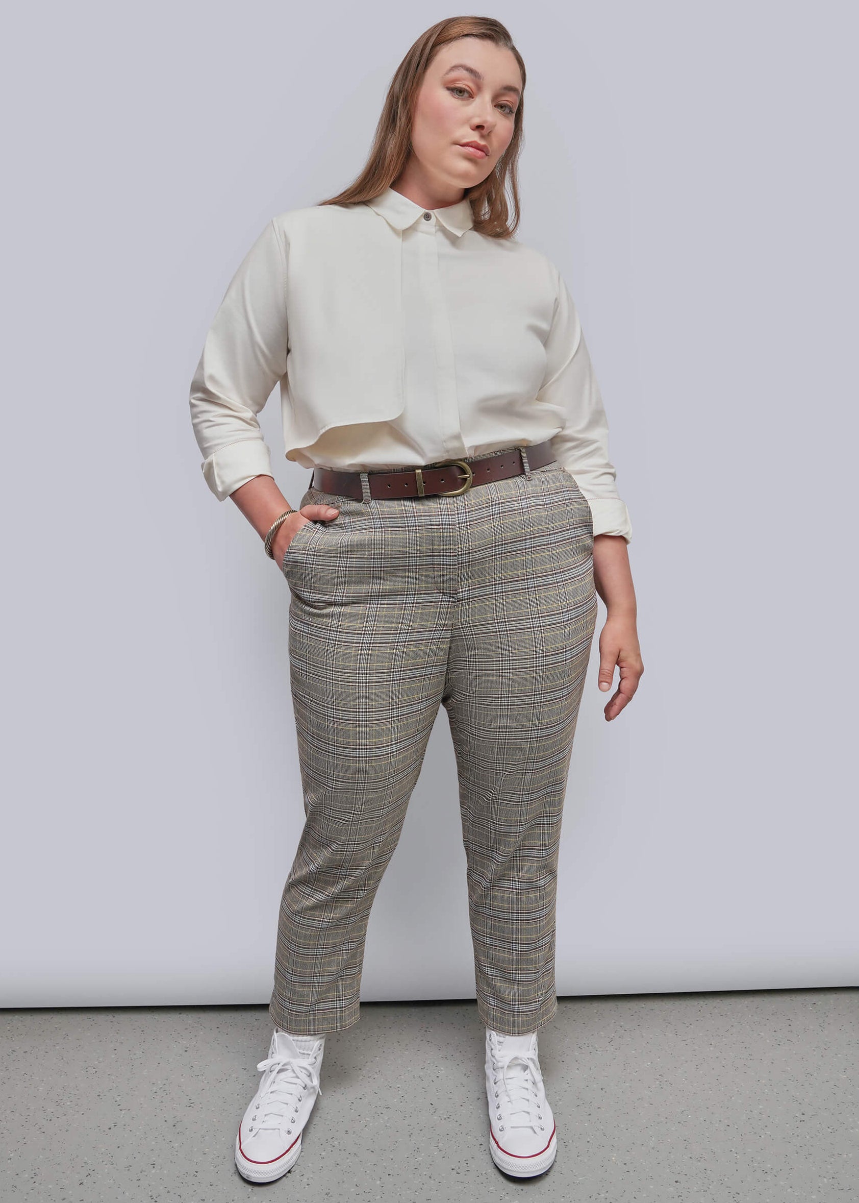 1X=A woman modeling a pair of beige and gray plaid trousers with a brown belt, paired with a cream shirt and white sneakers, standing against a light gray studio background.