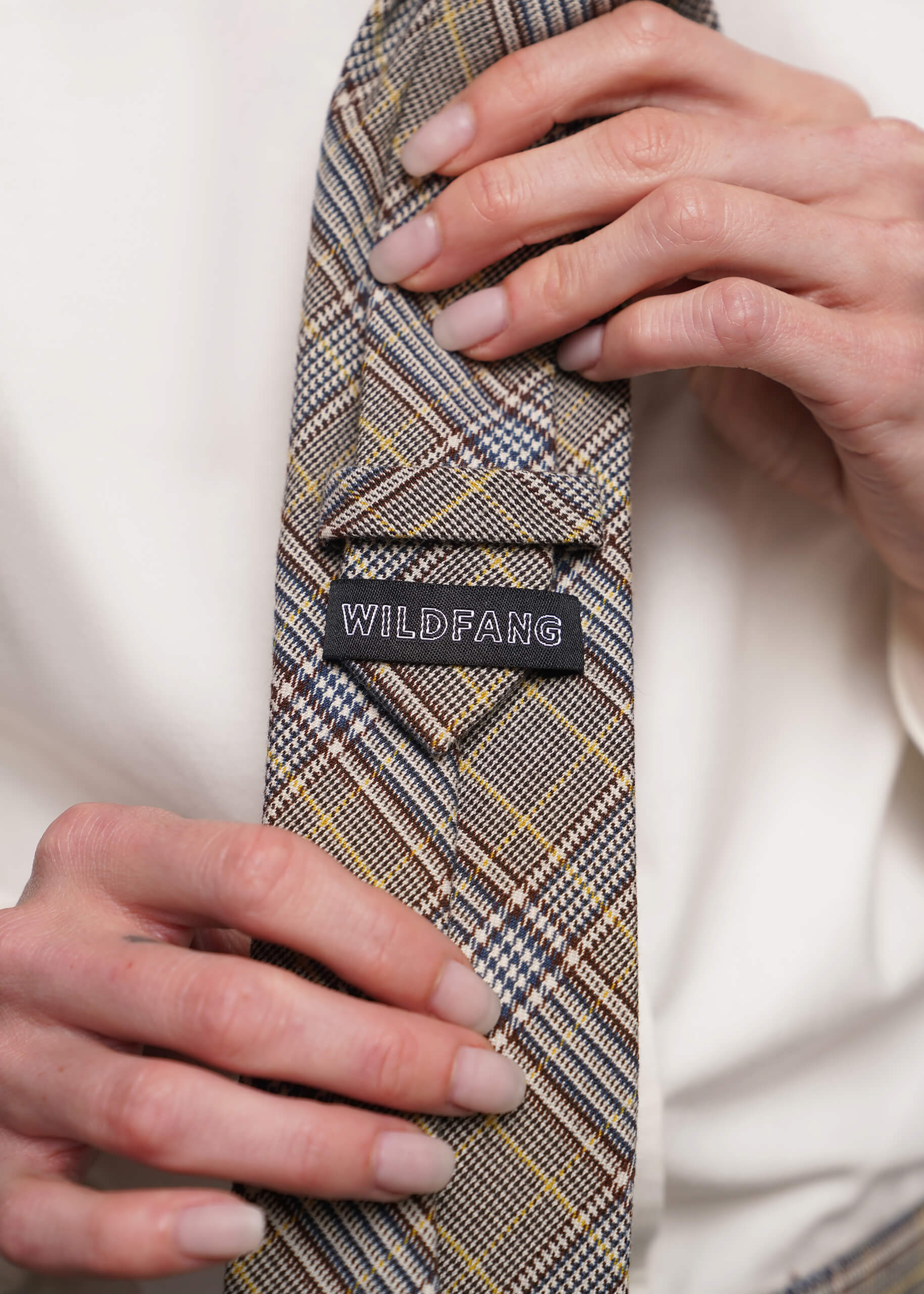 A close-up of a person's hands adjusting a plaid necktie with a black brand label that reads 'WILDFANG'. The tie features a beige, brown, and gray plaid pattern with thin yellow lines, worn with a white shirt.