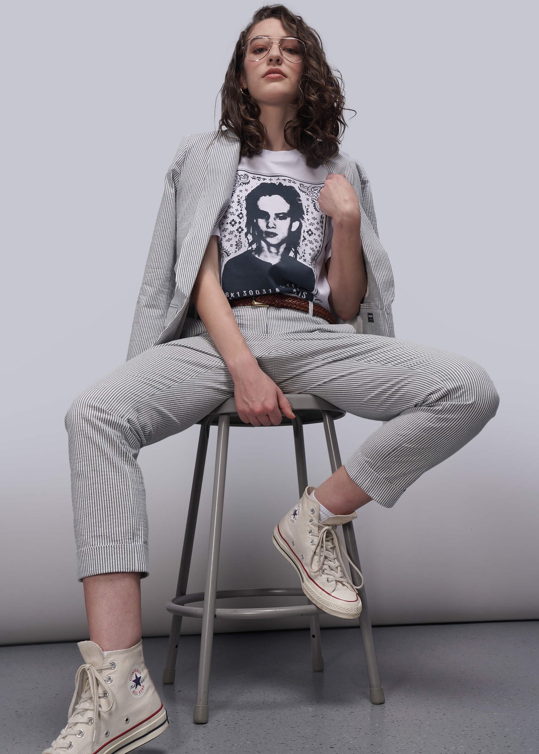 S=Woman sitting on a stool wearing a graphic t-shirt, light pants, and high-top sneakers against a plain background.