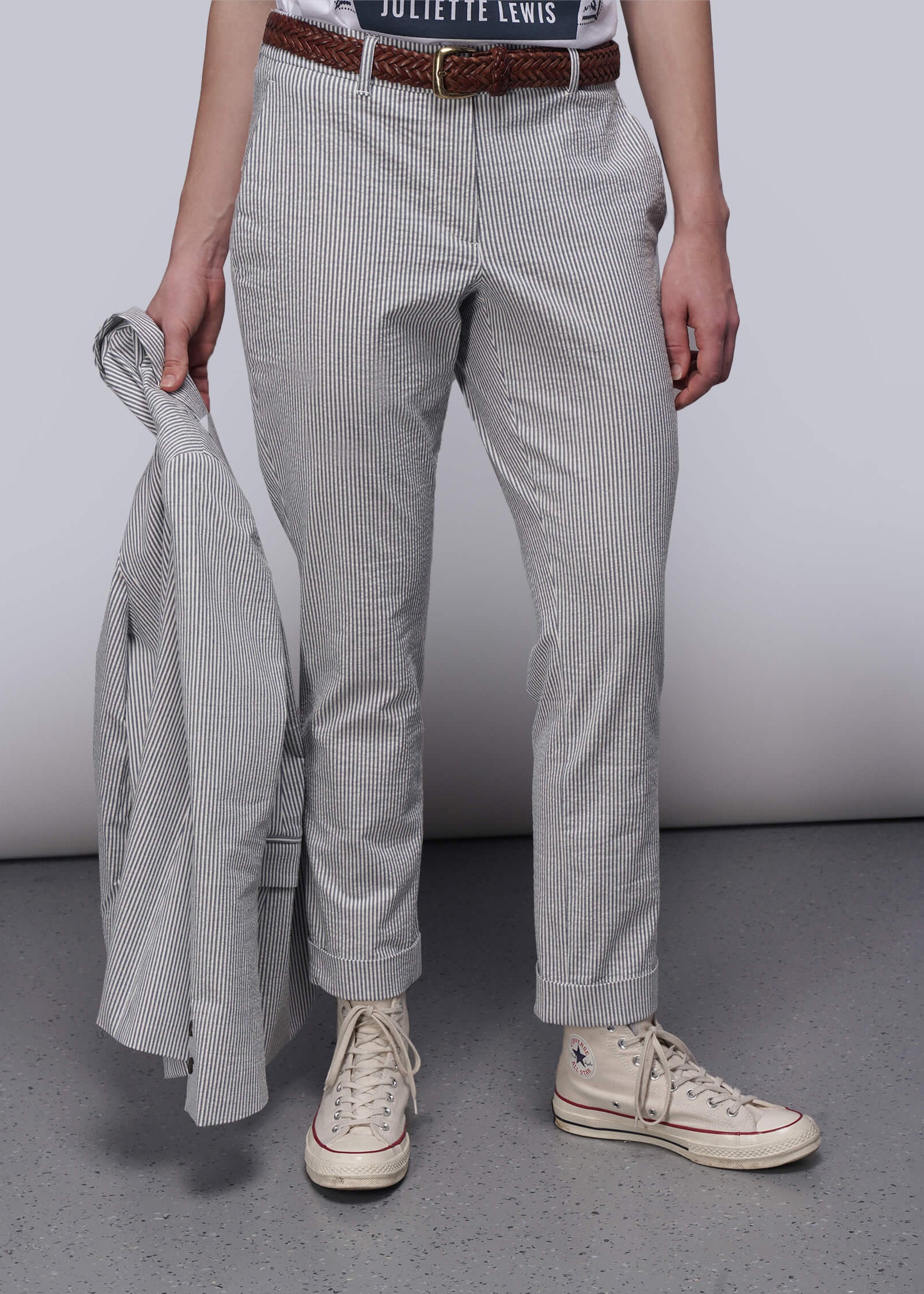Person wearing light blue striped pants and white sneakers holding a gray jacket against a plain background