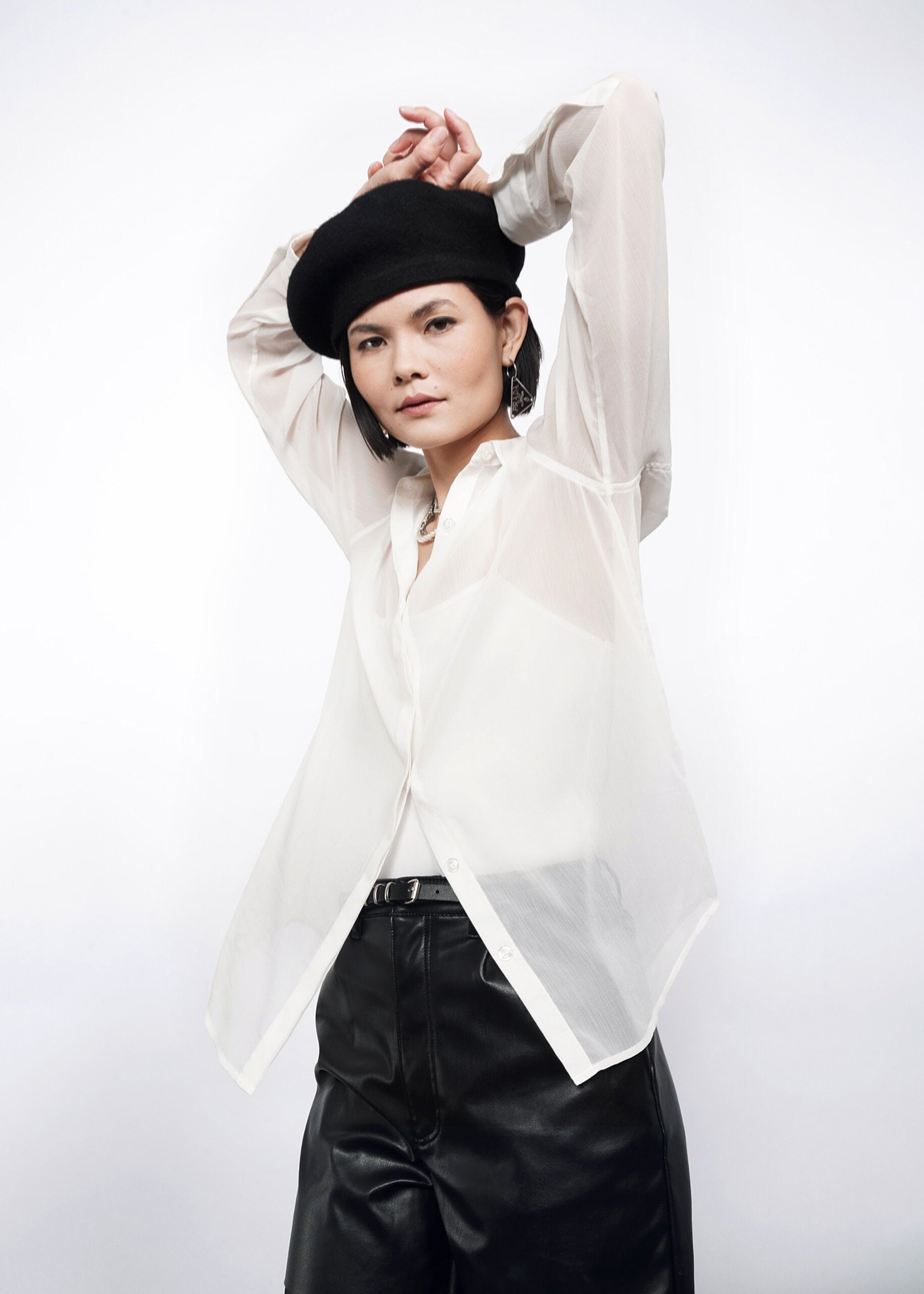 S=Someone poses with arms raised above their head in The Empower Sheer Long Sleeve Button Up—an oversized, flowy sheer top—paired with a black beret and black pants, against a plain white background.