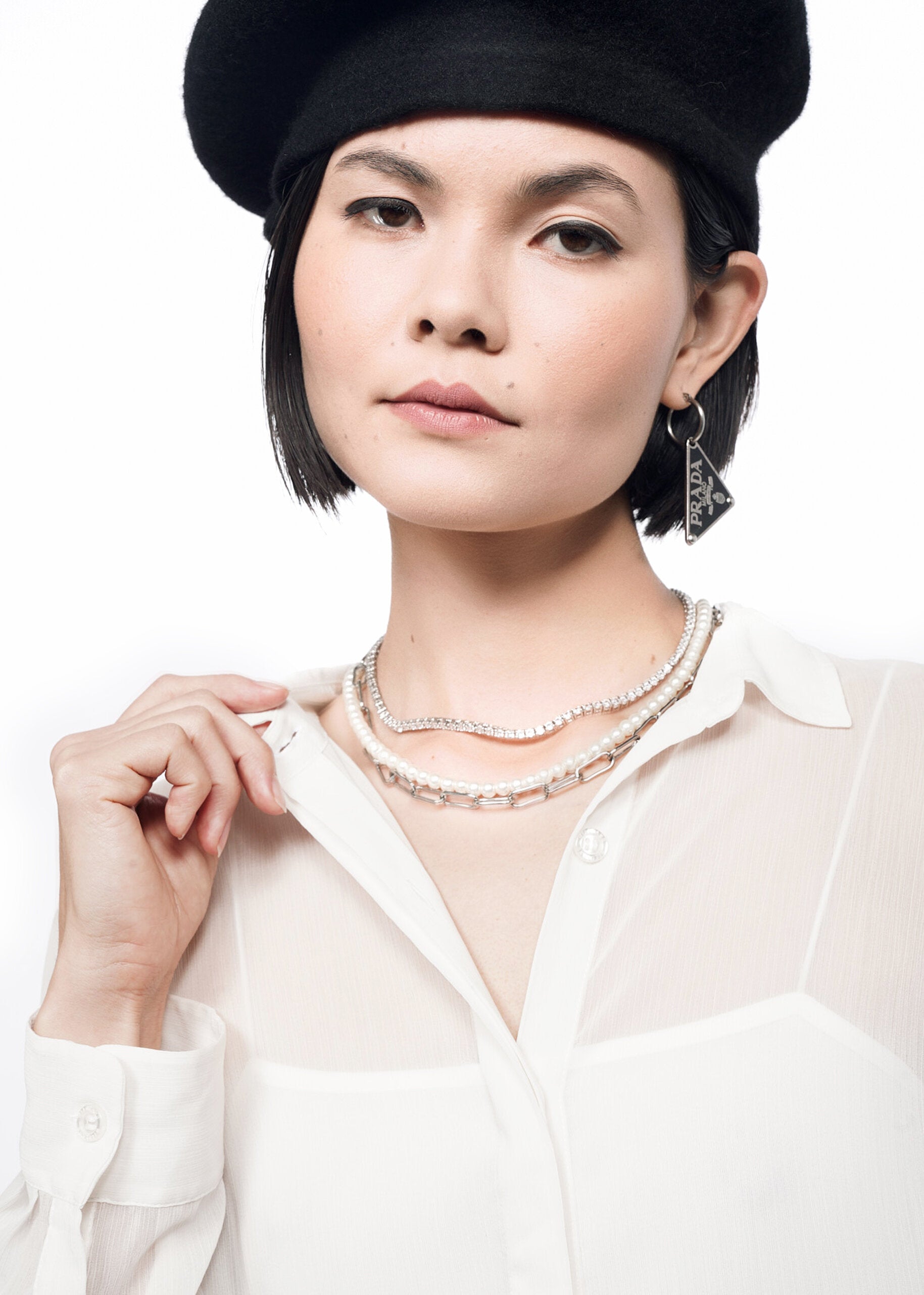 S= A woman confidently gazes at the camera while holding her collar, wearing The Empower Sheer Long Sleeve Button Up—a flowy top with Wildfang branded buttons—paired with light makeup, layered necklaces, and a single dangling earring.