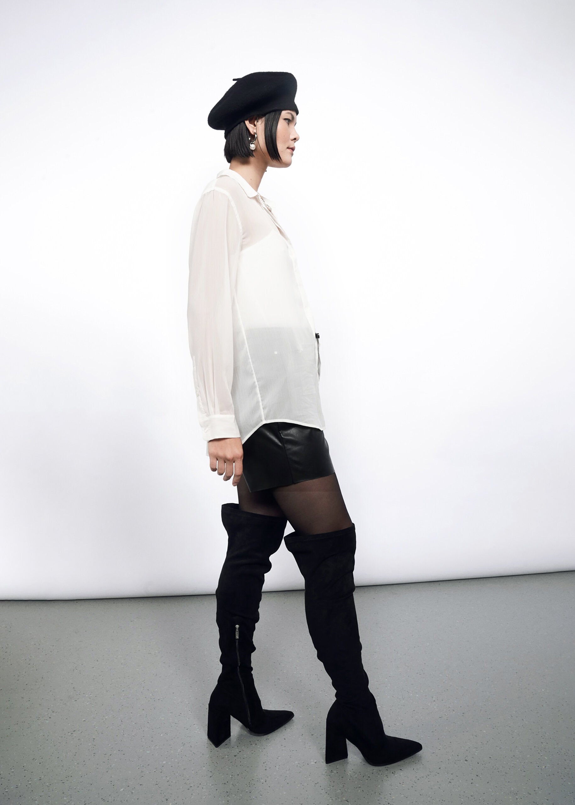 S=Wearing The Empower Sheer Long Sleeve Button Up, a person in a black beret, black mini skirt, and thigh-high black boots stands in profile against a plain white background.