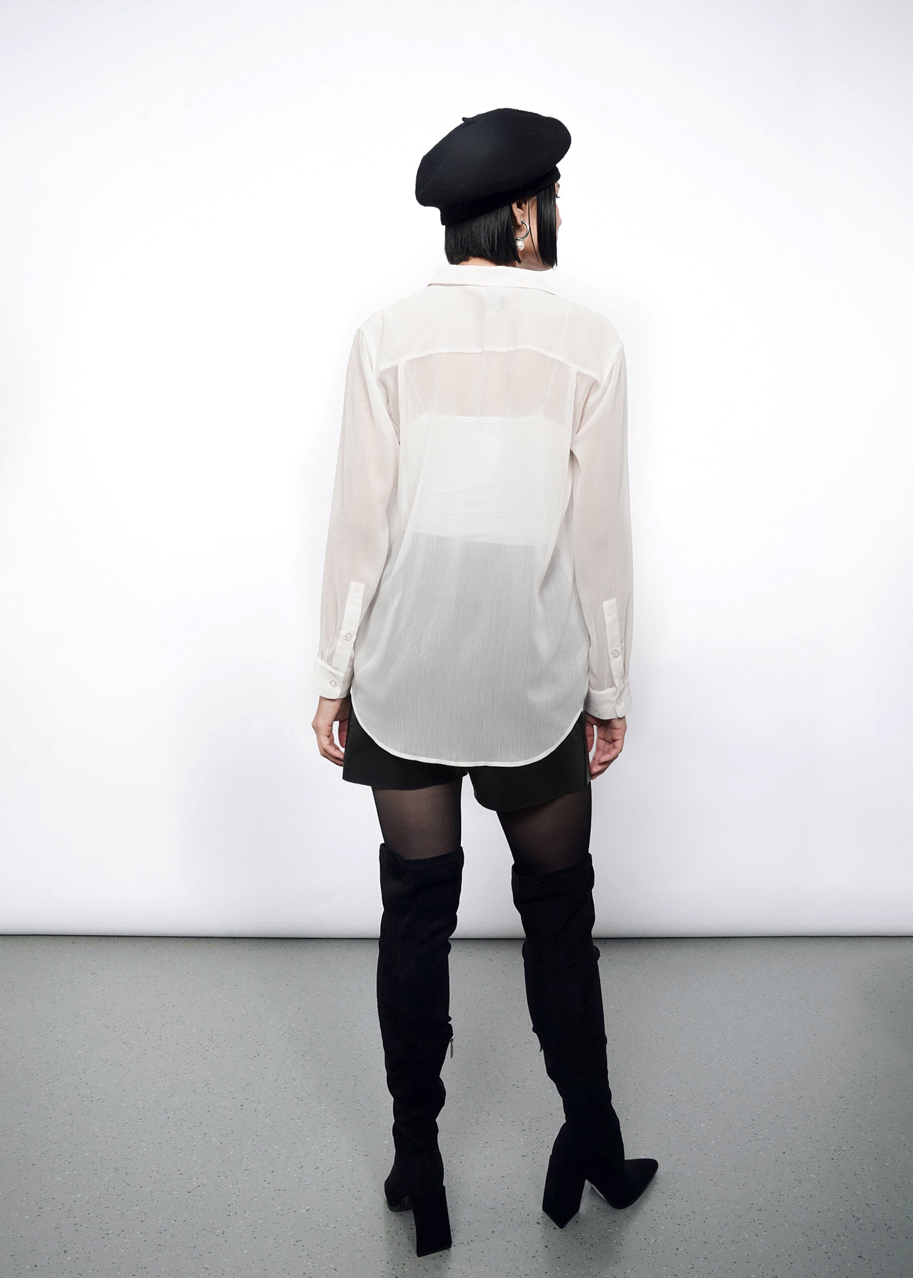S=A person faces away, wearing The Empower Sheer Long Sleeve Button Up with an oversized fit, a black beret, black shorts, and thigh-high boots. Background is a plain white wall with a gray floor.