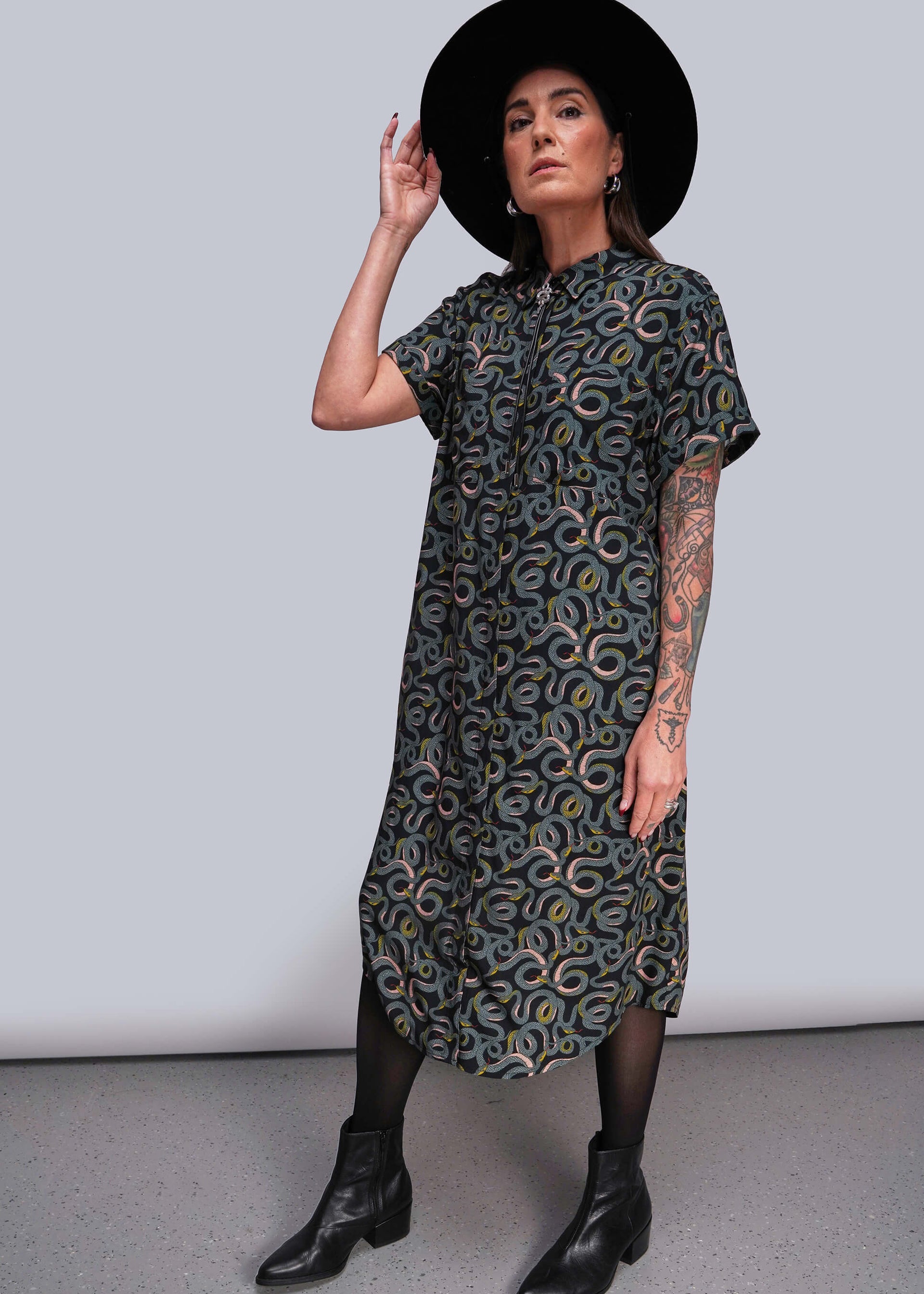 S=Woman wearing a patterned dress and black hat against a plain background