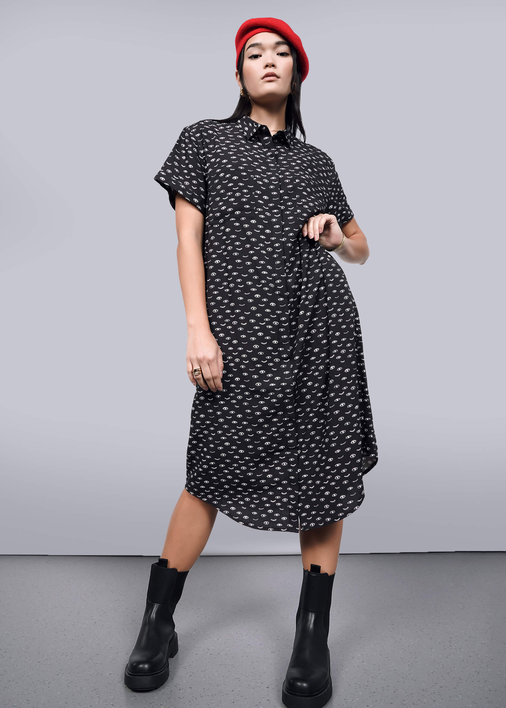 S= A woman in The Empower Shirt Dress, featuring a black pattern, button closure, and short sleeves below the knees, confidently poses with a red beret and black ankle boots. A plain background highlights her stylish outfit and poised stance.