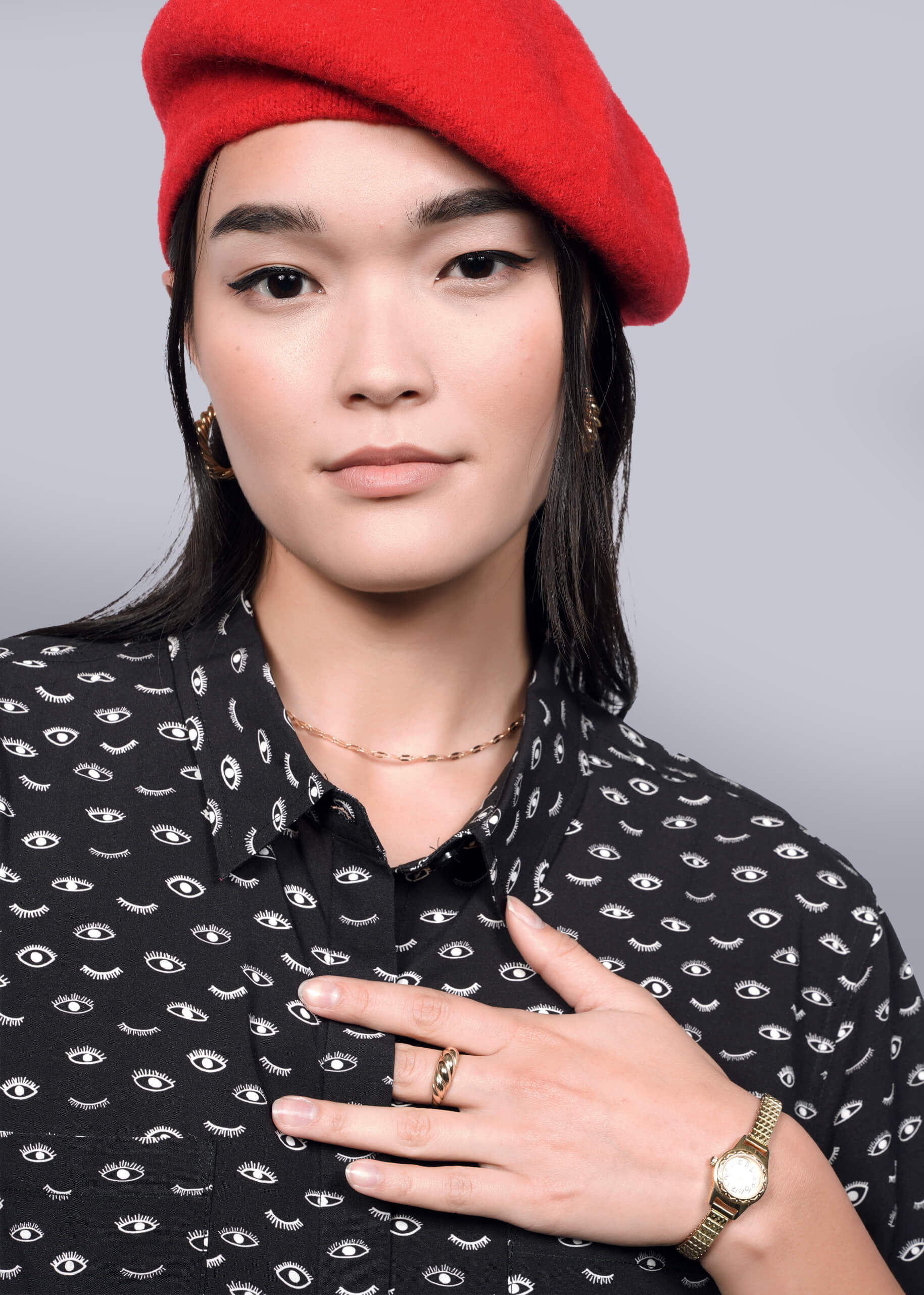 S= A person with long dark hair wears The Empower Shirt Dress, featuring eye patterns and a chic button closure, along with a red beret. They accessorize with gold hoop earrings, rings, and a watch. Their hand touches their chest against a white background.