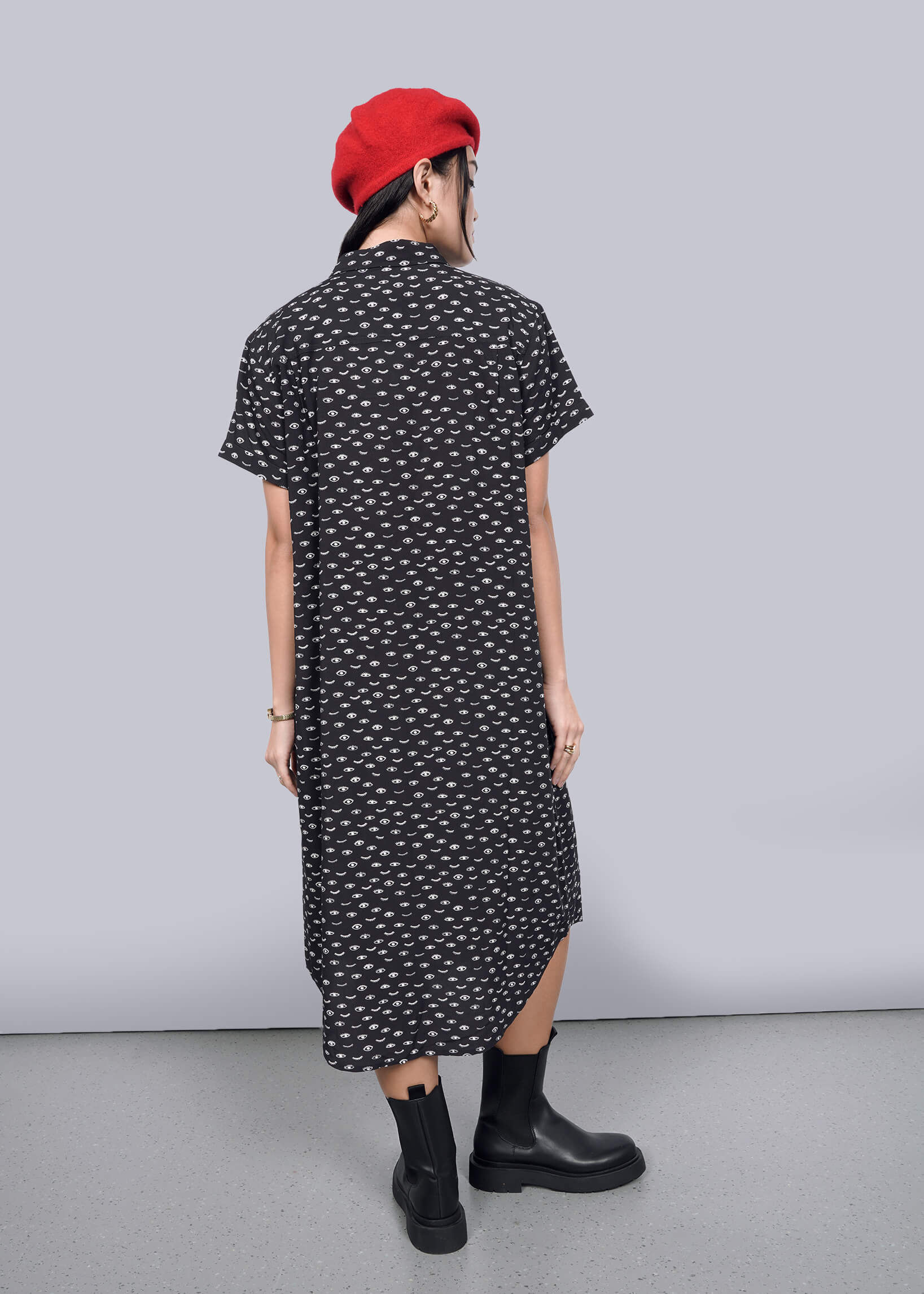 S=A person wearing The Empower Shirt Dress, with its black and white pattern, is paired with a red beret and black boots while standing against a plain white wall. Their hands rest by their sides on the light gray floor.