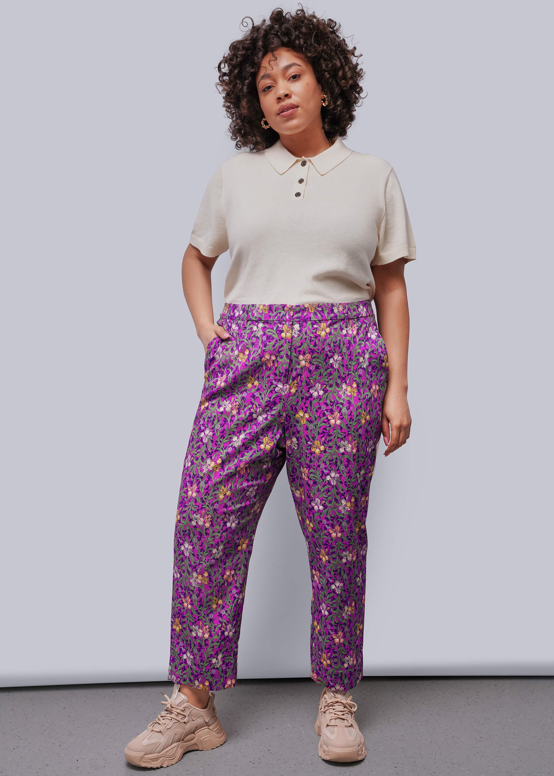 1X=Woman wearing a beige top and purple floral pants against a plain background