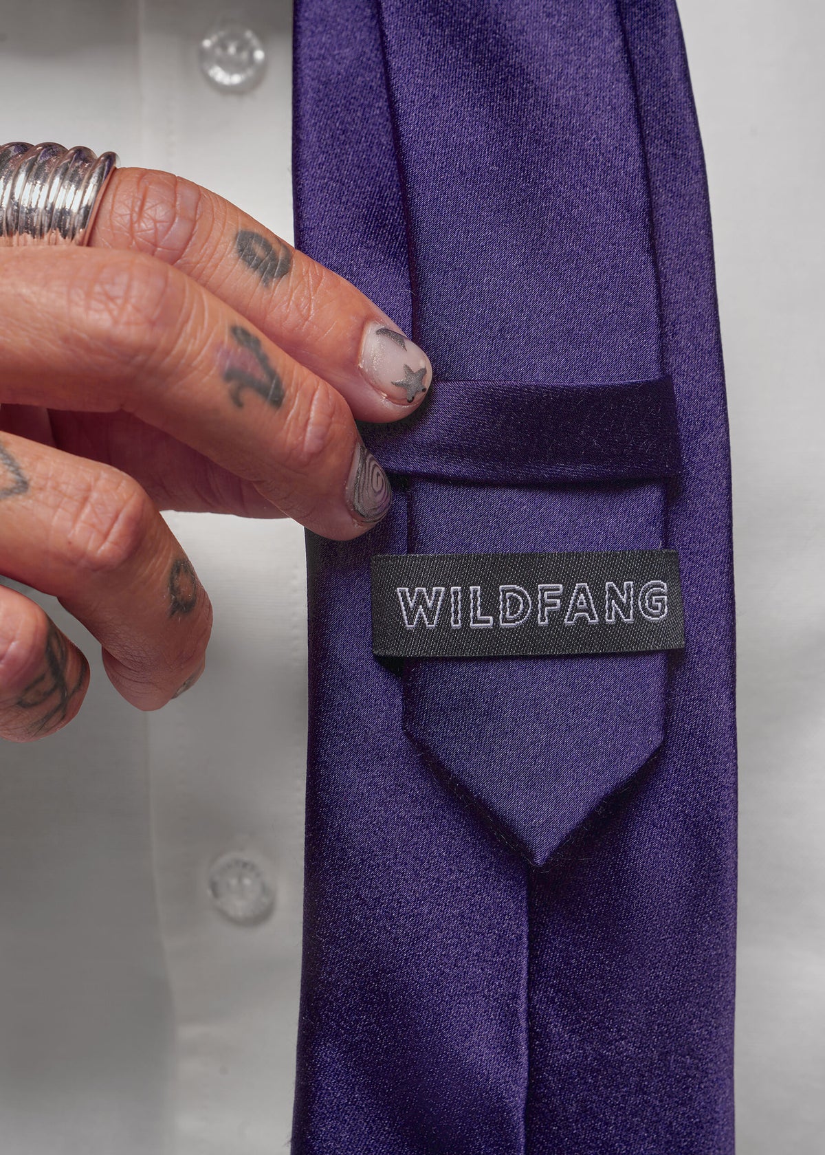 A close-up of a purple necktie with a black brand label reading 'WILDFANG', held by a tattooed hand against a white shirt background.