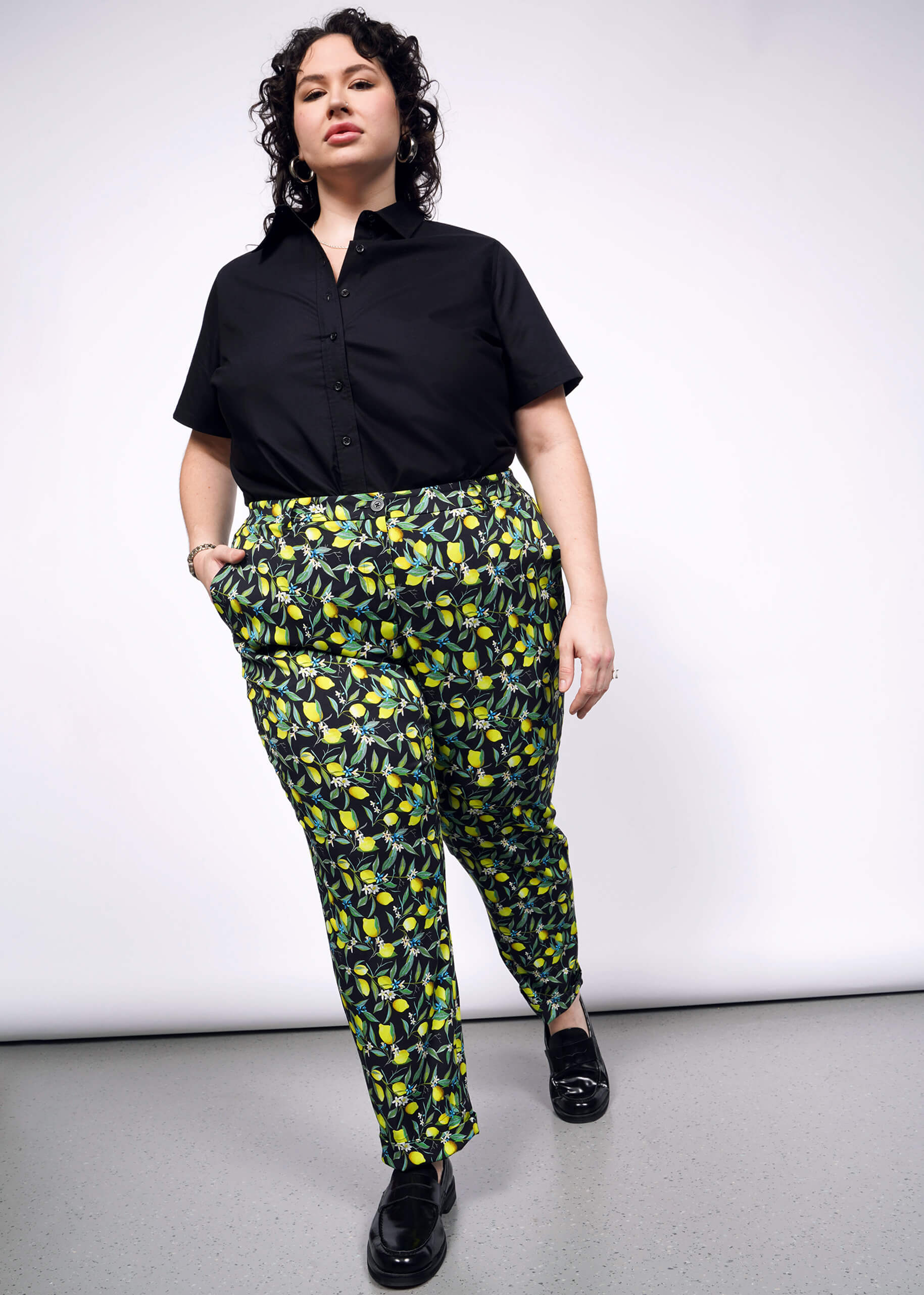 1X= Front full body view of person with curly short black hair wearing a black short sleeved button up and The Empower Trouser in Citrus Black, which is a yellow and green print on black background featuring lemons and leaves. They also wear black loafers.