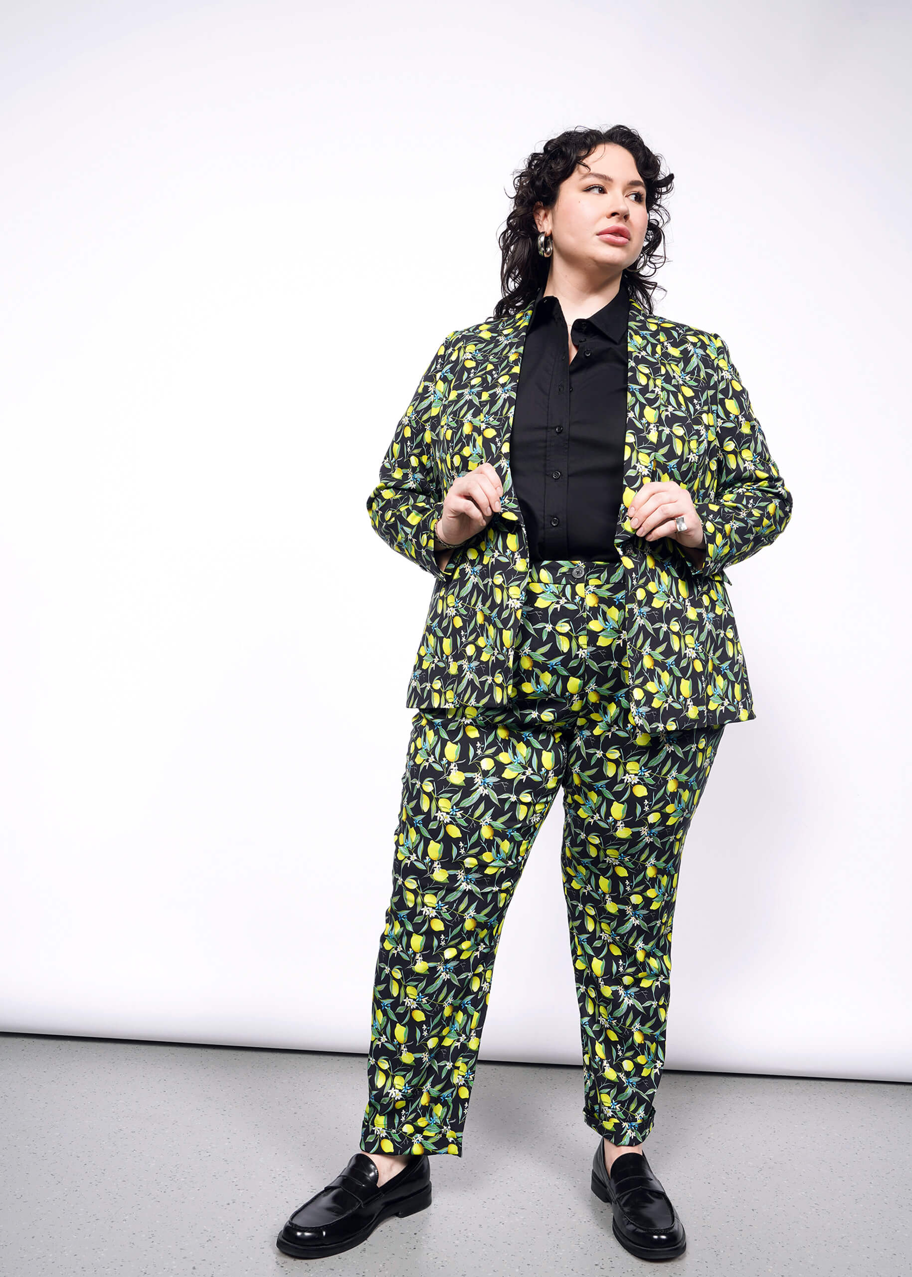 1X=Front full body view of person with curly short black hair wearing a black button up, with The Empower Tux Blazer and The Empower Trouser in Citrus Black, which is a yellow and green print on black background featuring lemons and leaves. They also wear black loafers.