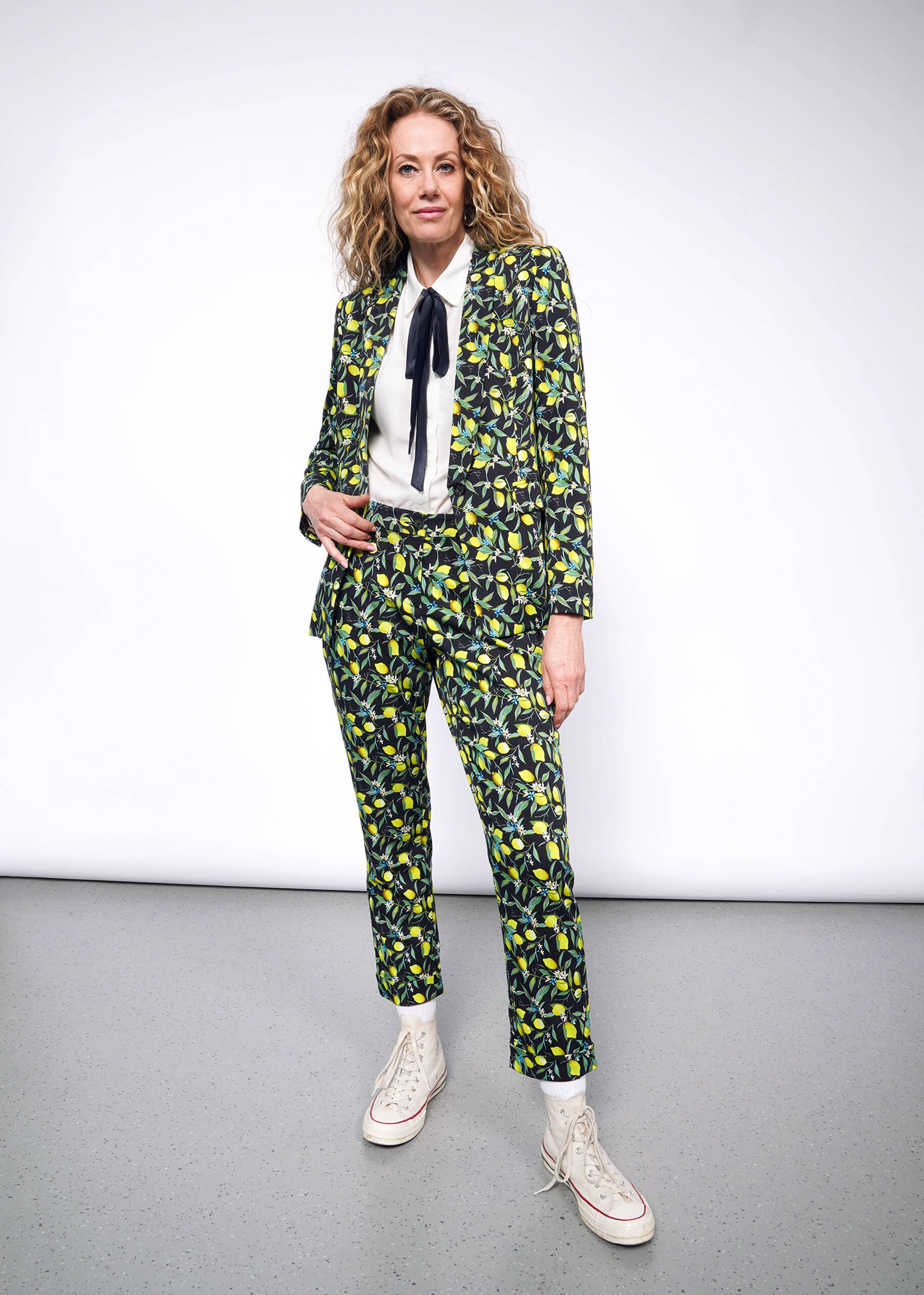 S=Front full body view of person with curly blonde hair wearing The Empower Bow Button Up in white, with The Empower Tux Blazer and The Empower Trouser in Citrus Black, which is a yellow and green print on black background featuring lemons and leaves. They also wear white Converse sneakers.