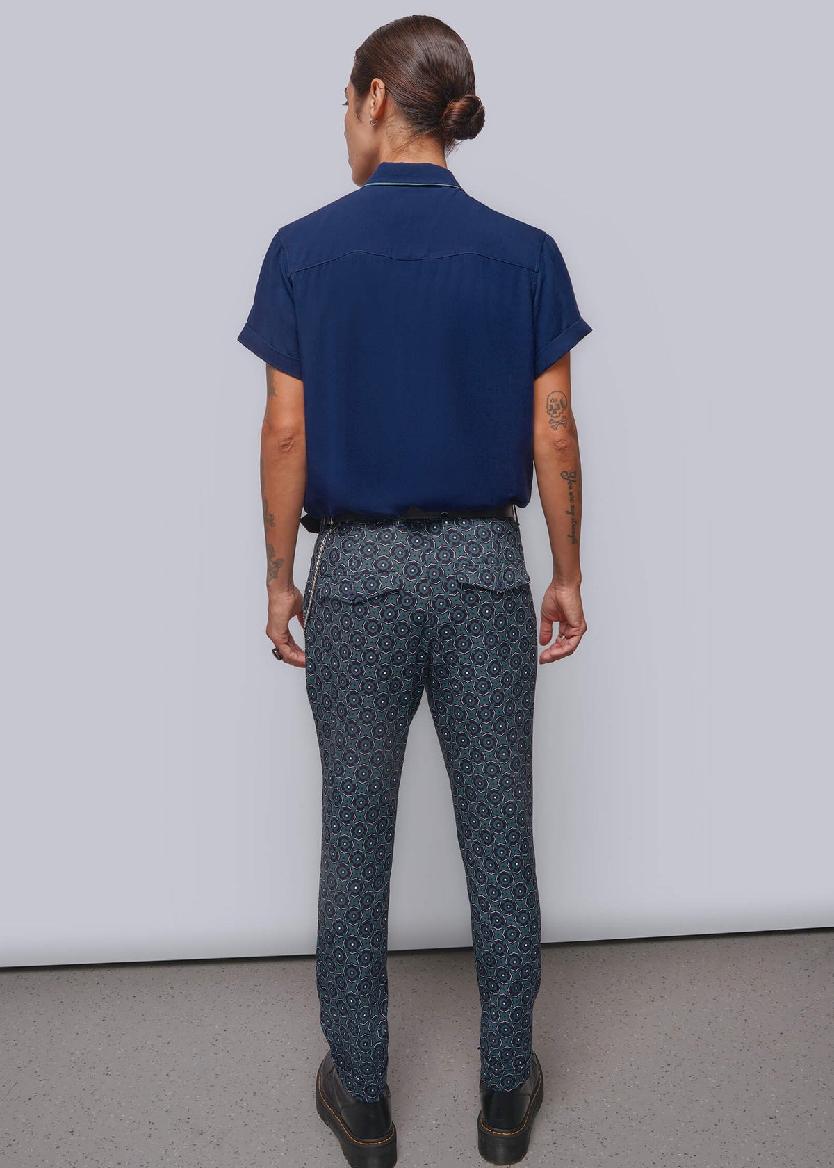 S=A model is shown from the back wearing a navy blue short-sleeve collared shirt and dark blue patterned trousers, standing against a light gray studio background.