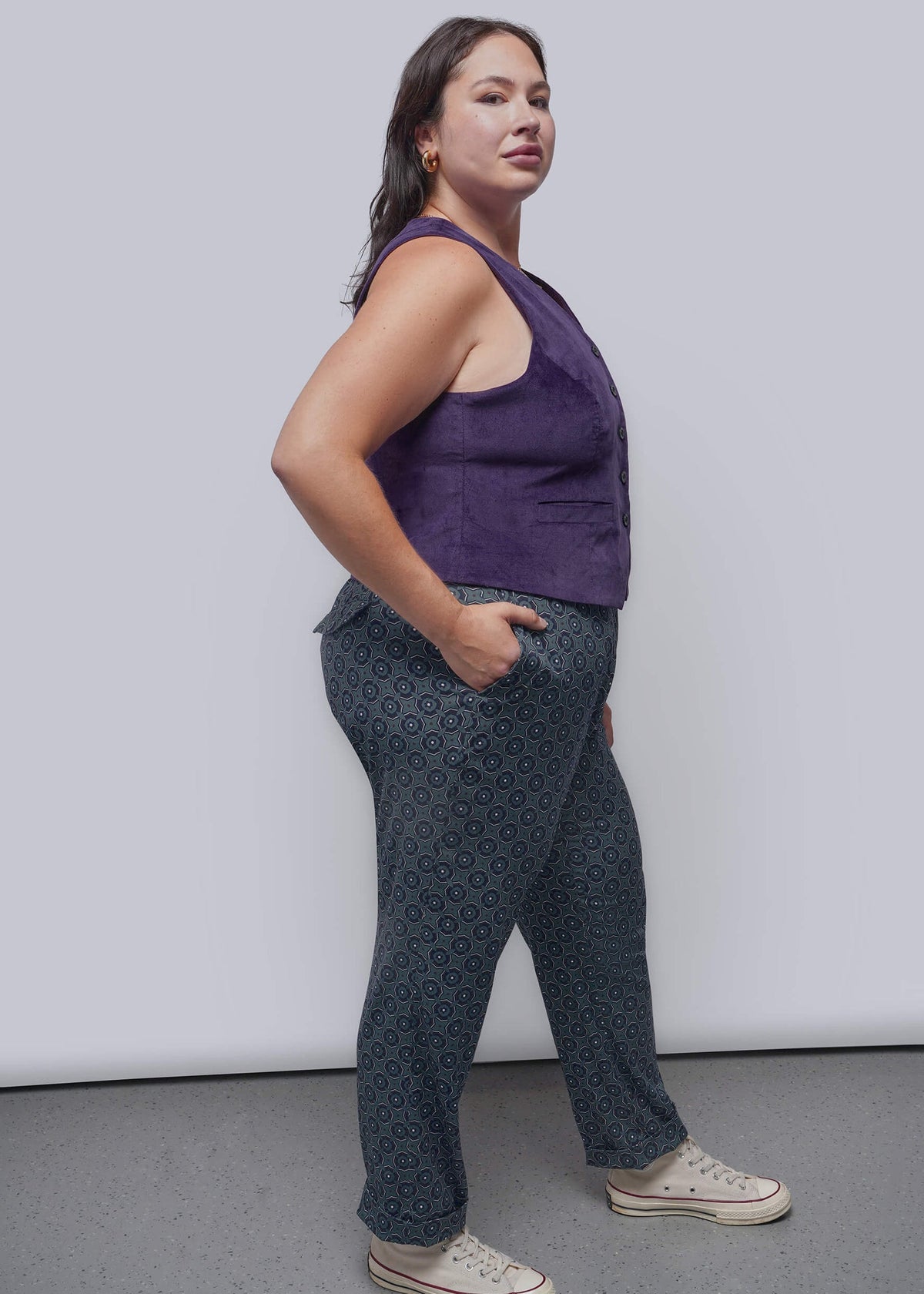 1X=A woman modeling a sleeveless purple top and dark patterned pants with white sneakers, standing against a light gray studio background.