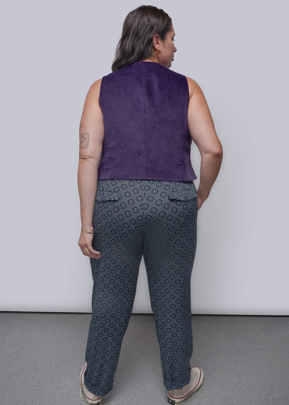 1X=A woman is shown from behind wearing a sleeveless purple vest and dark gray patterned trousers with beige shoes, standing against a plain light gray wall.