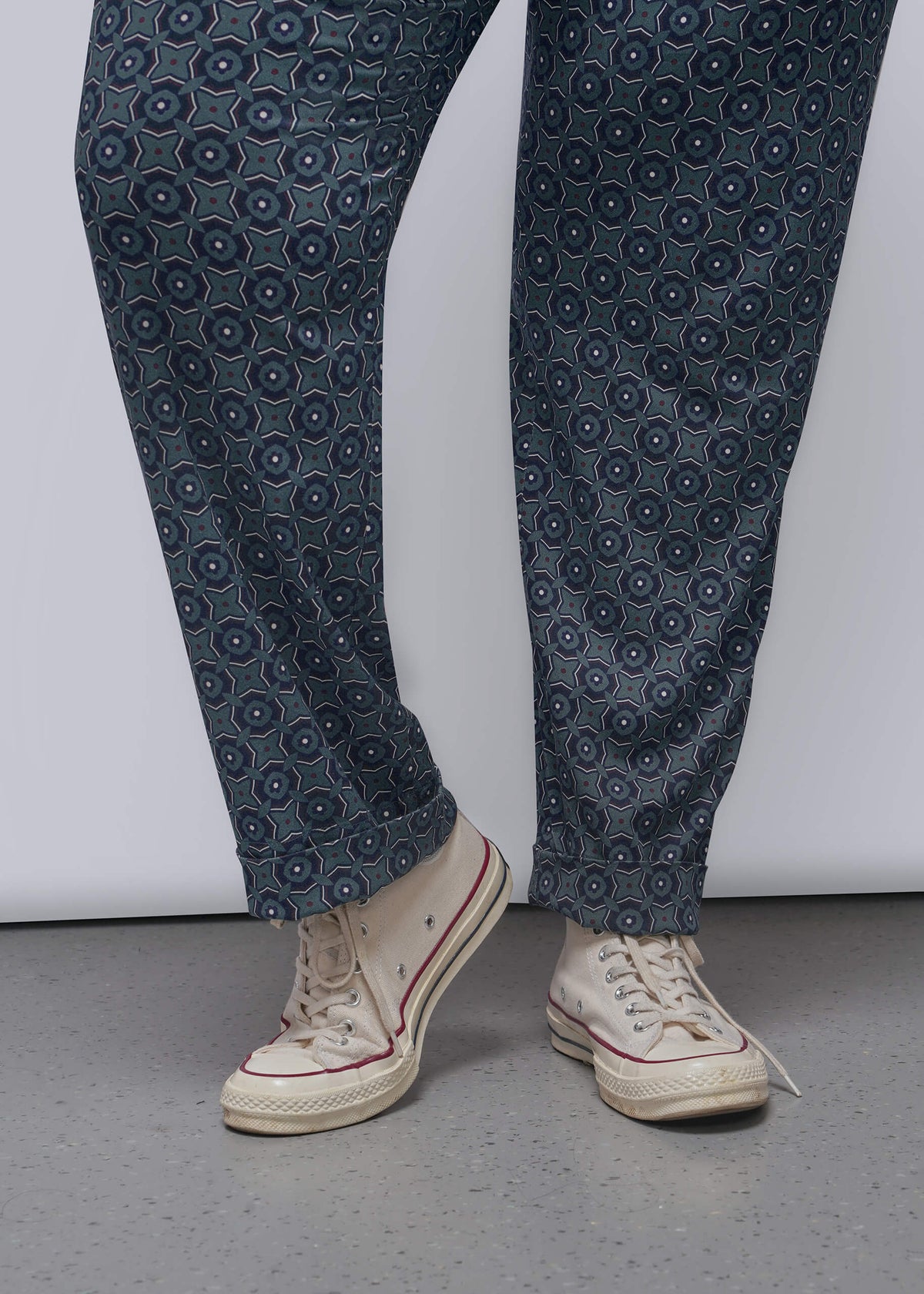 1X=A person wearing dark blue patterned trousers and cream high-top sneakers, standing against a light gray wall and concrete floor.