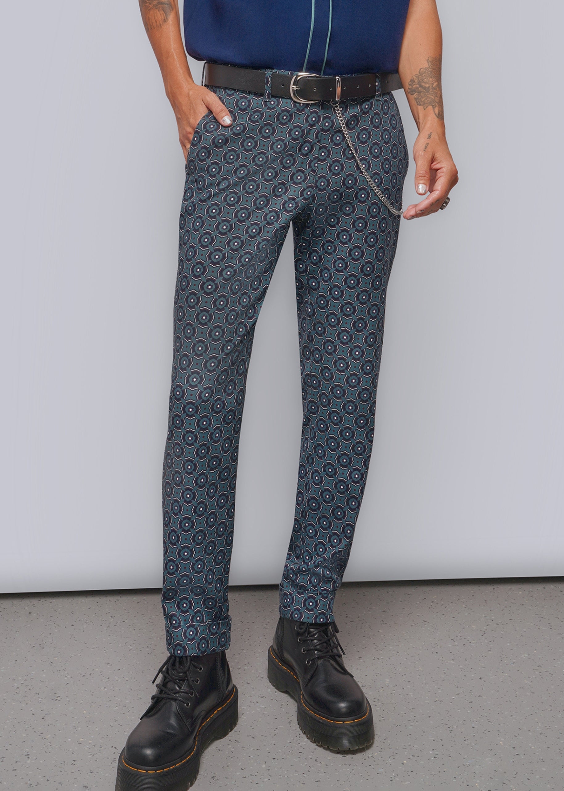 A person wearing trousers with a repeating circular geometric pattern in dark blue, gray, and black, styled with a black belt, chain accessory, navy top, and black boots against a plain light gray studio background.