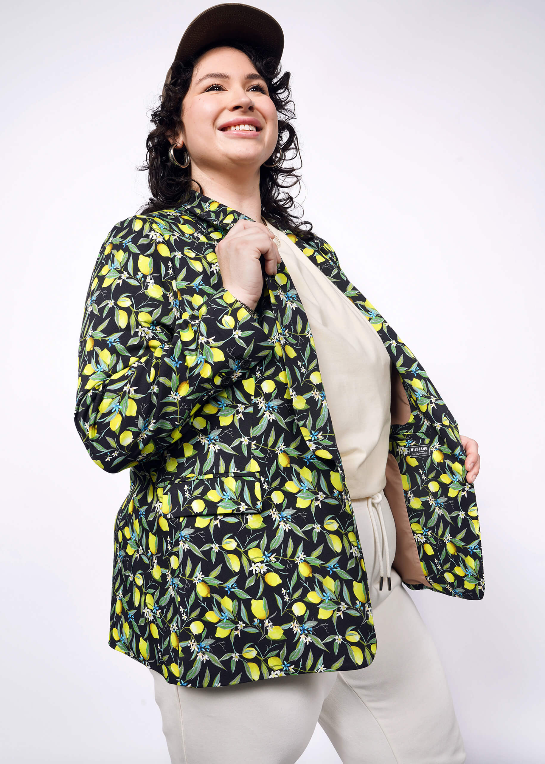 1X=Person with short curly black hair wear black cap, tan colored shirt and pants, with The Empower Tux Blazer in Citrus Black, which is a yellow and green print on black background featuring lemons and leaves.