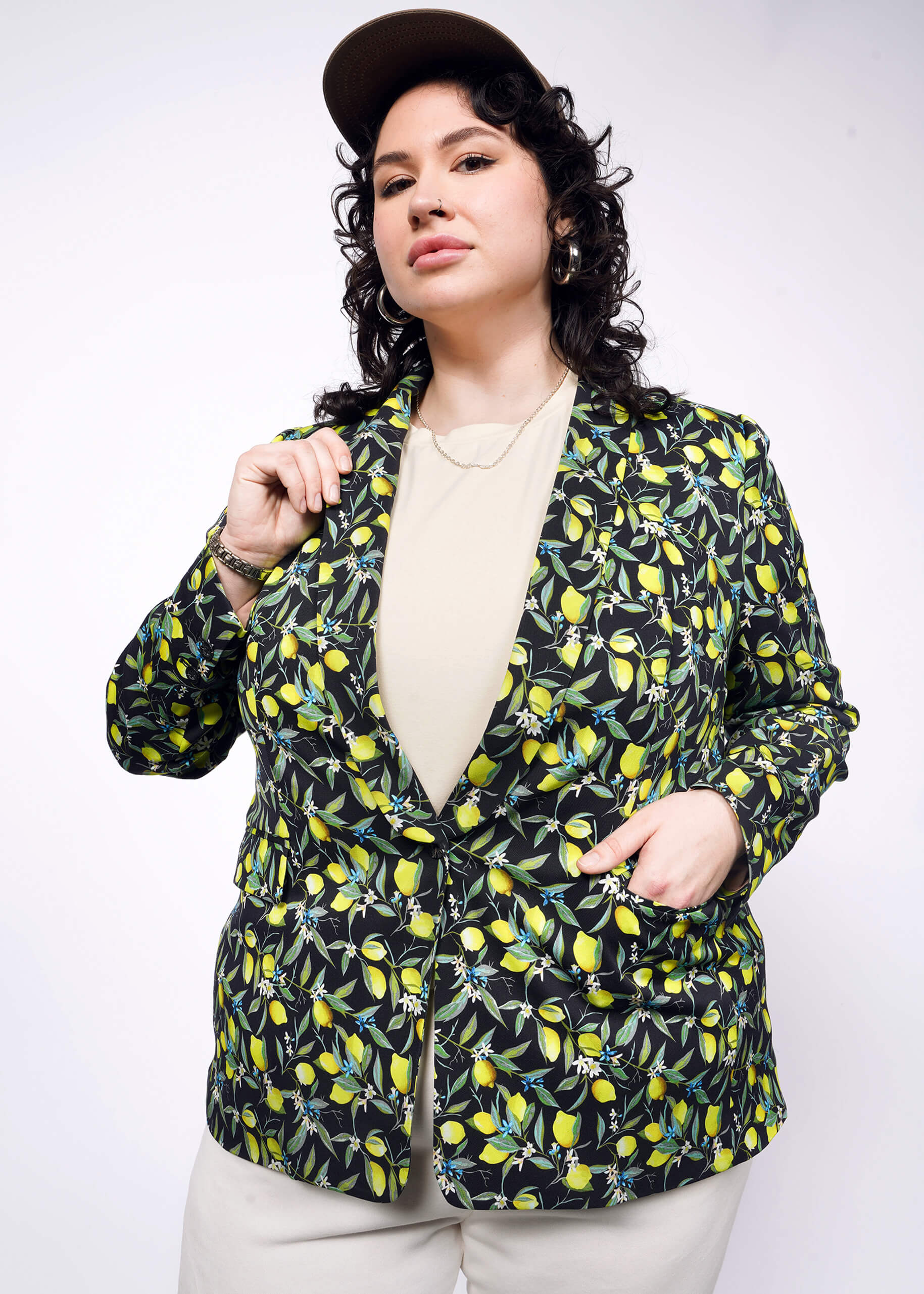1X= Front view of person with short curly black hair wearing black cap, tan colored shirt and pants, with The Empower Tux Blazer in Citrus Black, which is a yellow and green print on black background featuring lemons and leaves. 