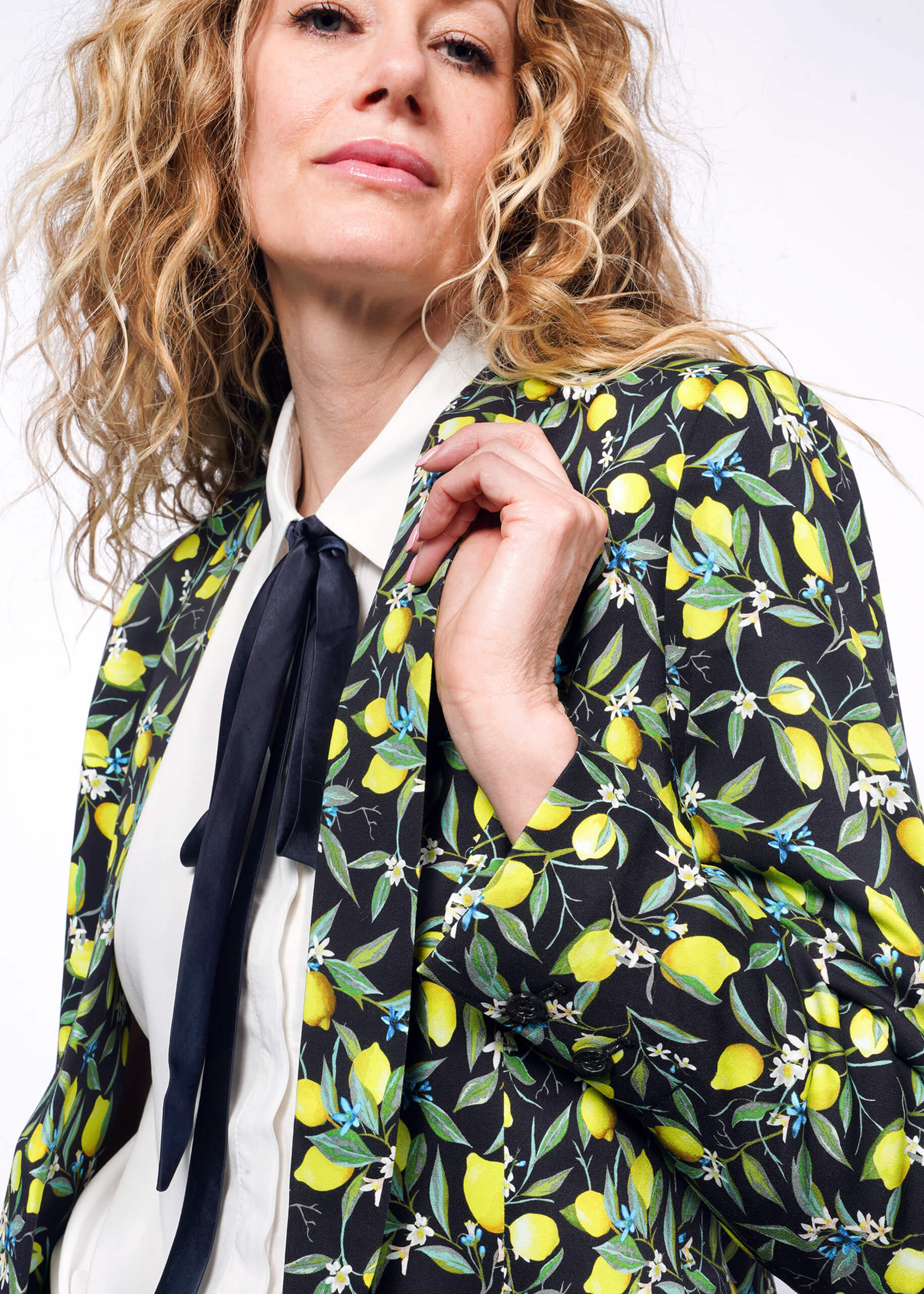 S=Close up side view of person with curly blonde hair wearing The Empower Bow Button Up in white, with The Empower Tux Blazer in Citrus Black, which is a yellow and green print on black background featuring lemons and leaves.