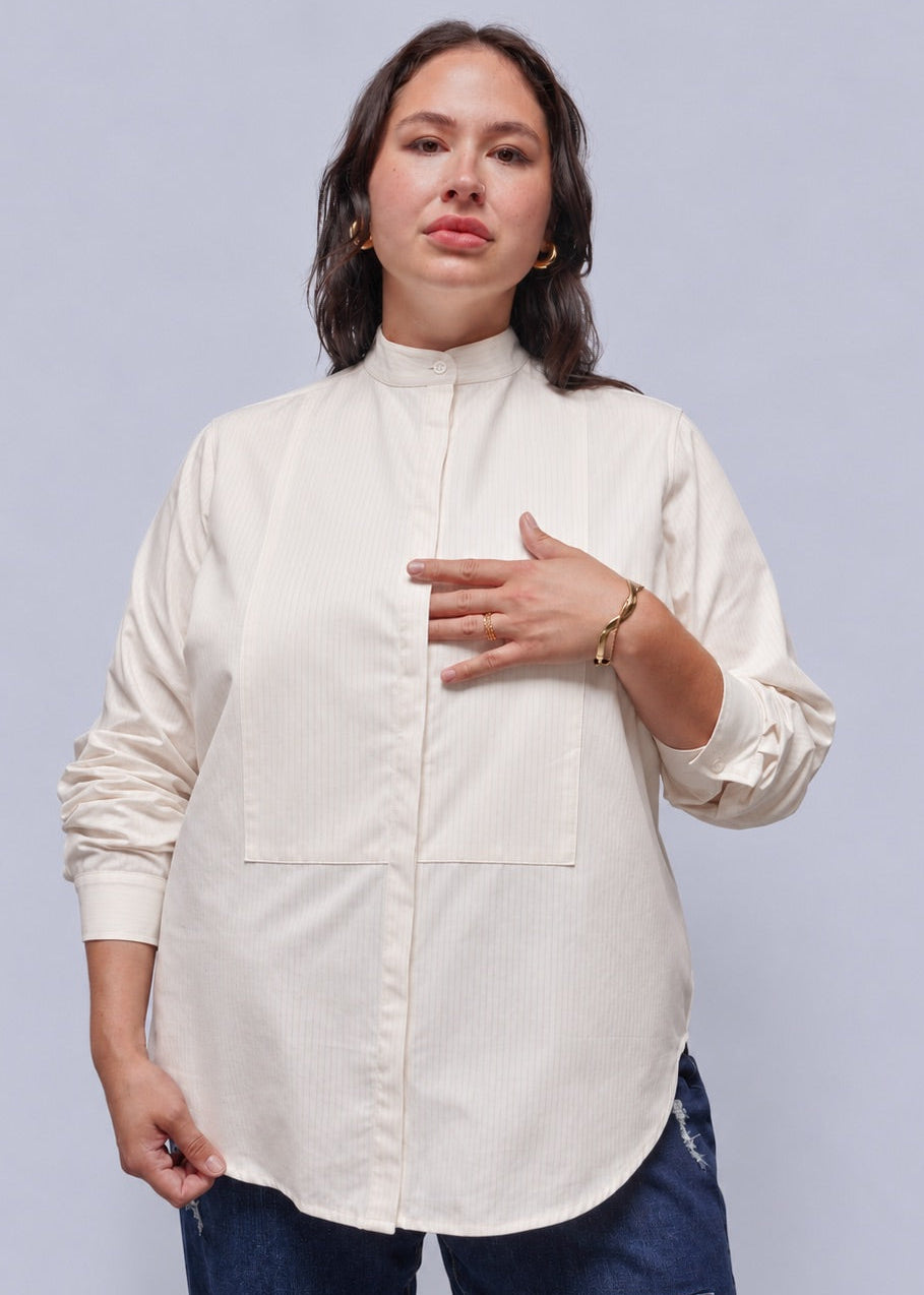 1X=A woman modeling a high-neck, long-sleeve blouse in a light cream or off-white color, paired with dark blue jeans, against a solid light gray studio background.