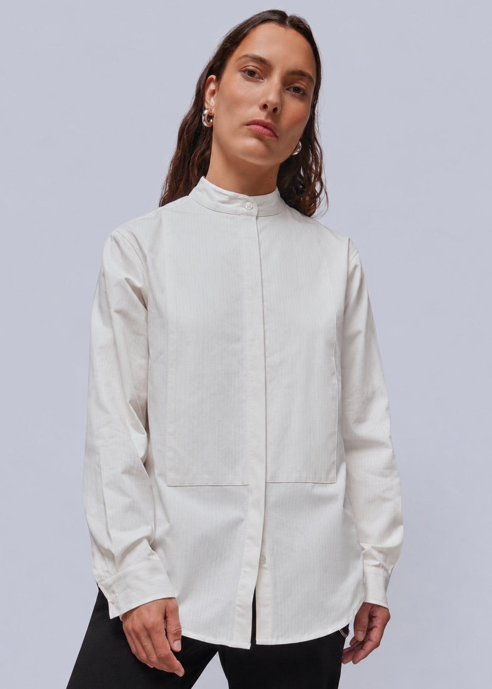 S=A woman modeling a high-neck, long-sleeve white button up, paired with black pants, against a solid light gray studio background.