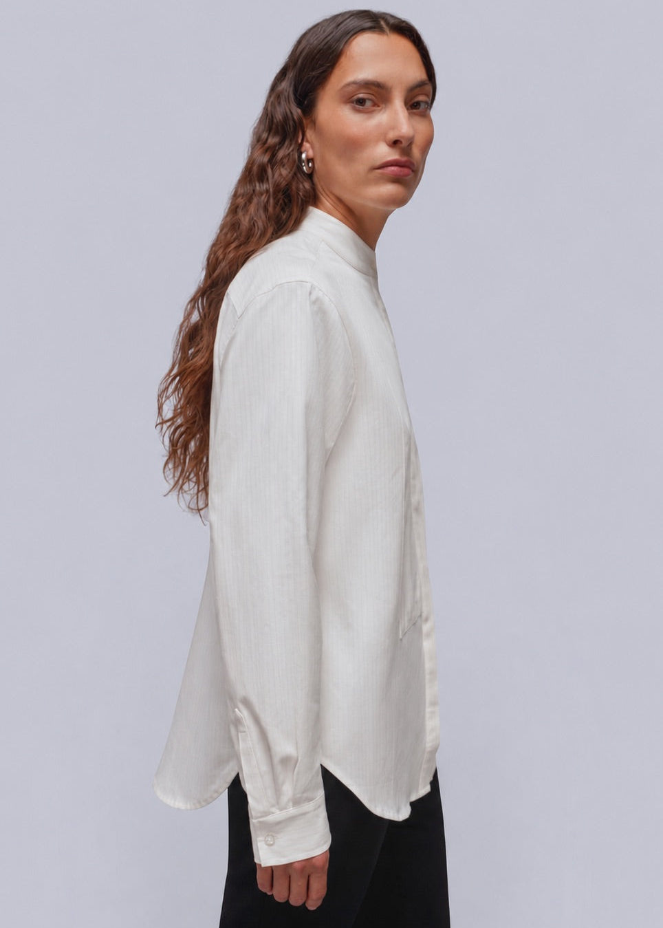 S=A woman with long brown hair is shown in profile wearing a white long-sleeve button-up shirt and dark pants against a light gray studio background.