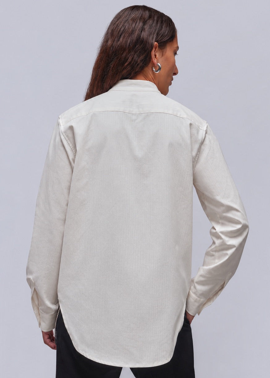 S=A woman shown from the back wearing a light cream-colored long-sleeve shirt and black pants, set against a plain light gray studio background.