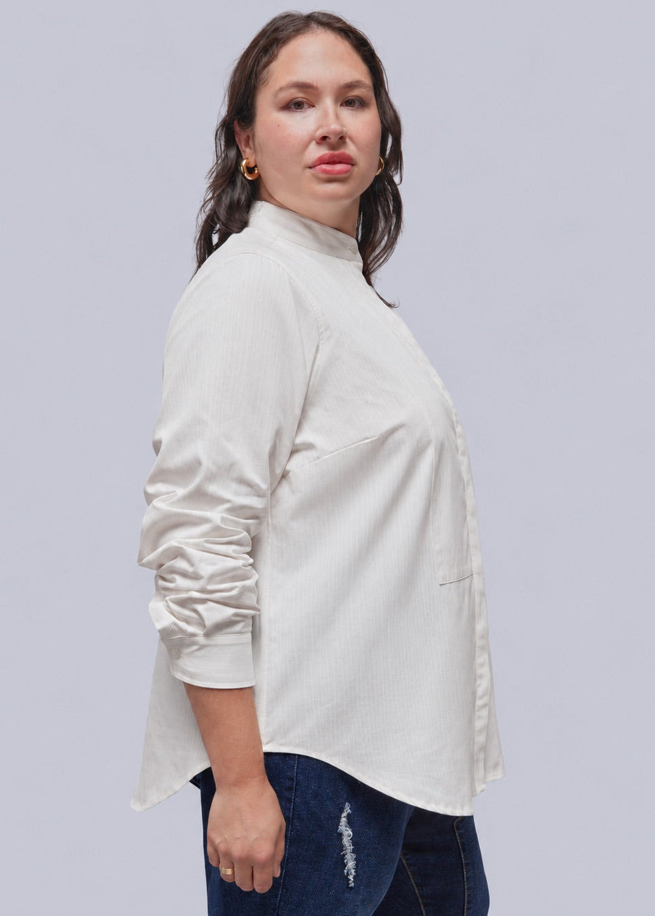 1X=A woman modeling a high-neck, long-sleeve button-up blouse in a light cream or off-white color, paired with dark blue jeans, against a solid light gray background.