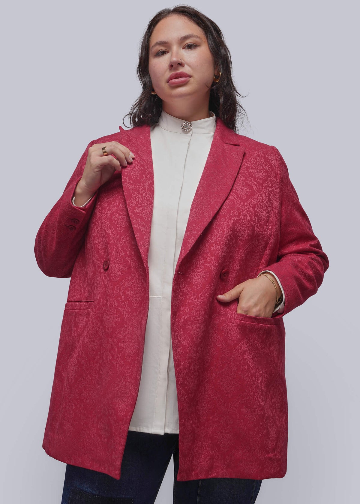 1X=A woman modeling a textured, double-breasted blazer in a deep magenta pink, worn over a cream mock-neck top and dark blue jeans, against a solid light gray background.