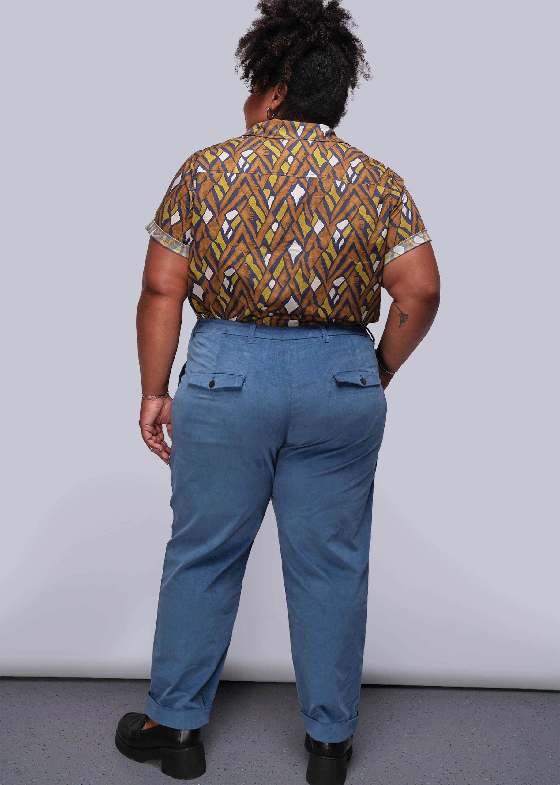 1X=Person wearing a patterned shirt and blue pants against a plain background