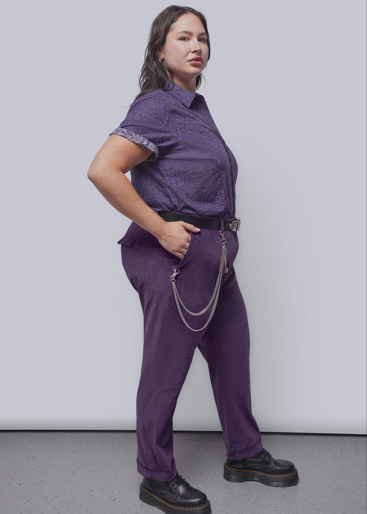 1X=A woman modeling a purple short-sleeve patterned button-up shirt and matching purple trousers, styled with a black belt, silver chain accessory, and black platform shoes, against a light gray studio background.
